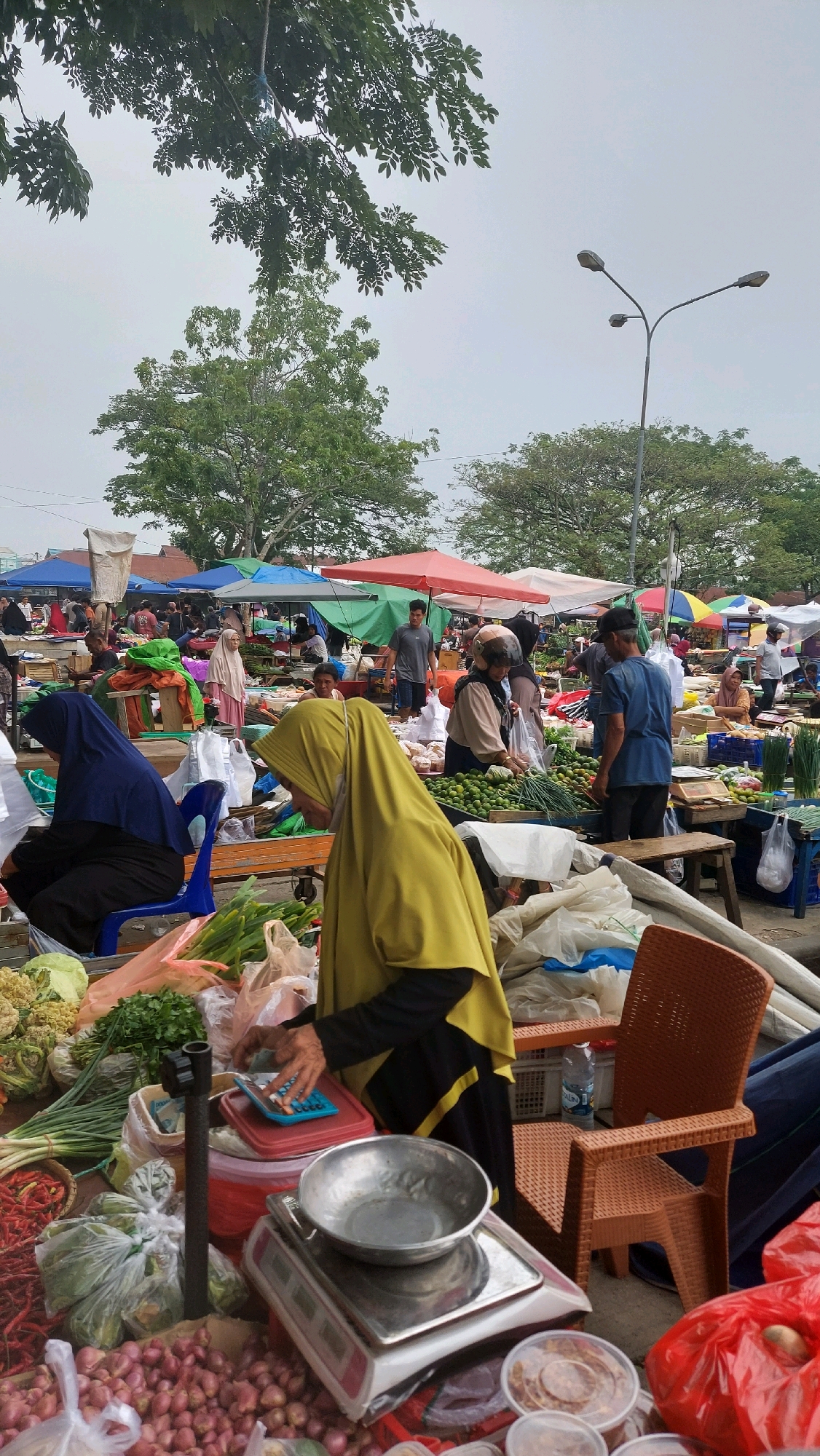 In Sungai Bedungun, Indonesia, a bustling outdoor market is captured under an overcast sky. The scene is filled with a variety of produce, including red chilies, onions, cauliflower, and leafy green vegetables, displayed on wooden tables and in plastic bags. Several people are present, some of whom appear to be vendors and customers. One woman, wearing a yellow hijab and black and yellow garment, is seen operating a blue calculator, with a silver weighing scale and a metal bowl in front of her. Another woman, in a blue hijab, is seated on a blue chair. Other individuals in the background are browsing or interacting with stalls, some of which are covered by colorful awnings. The environment suggests a typical market day with commercial activity.