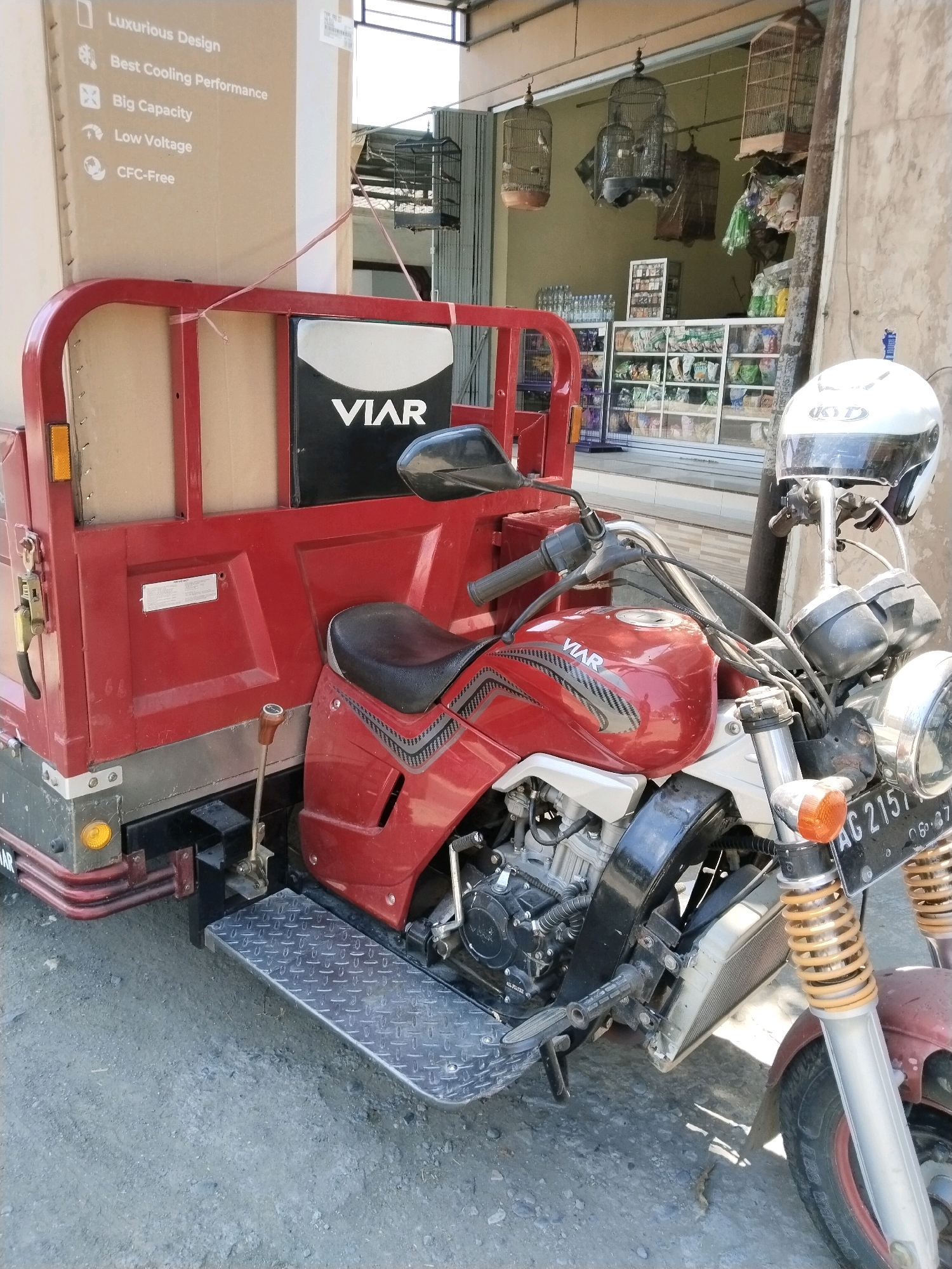 The image shows a red three-wheeled motorcycle with a cargo box attached, parked on a gravel surface in front of a store. The cargo box is large and appears to be made of cardboard, with text in Indonesian suggesting it contains a product with "Luxurious Design," "Best Cooling Performance," "Big Capacity," "Low Voltage," and is "CFC-Free." The store behind the motorcycle has shelves stocked with various items, and several birdcages hang from the ceiling. A white helmet with a black visor is placed on the handlebars of the motorcycle. The license plate of the motorcycle reads "AG 2157." The environment appears to be an outdoor market or a commercial street in Mergayu, Indonesia, given the architecture and the presence of goods typically sold in such locations. The lighting suggests it is daytime, with direct sunlight casting shadows. There are no people visible in the image.