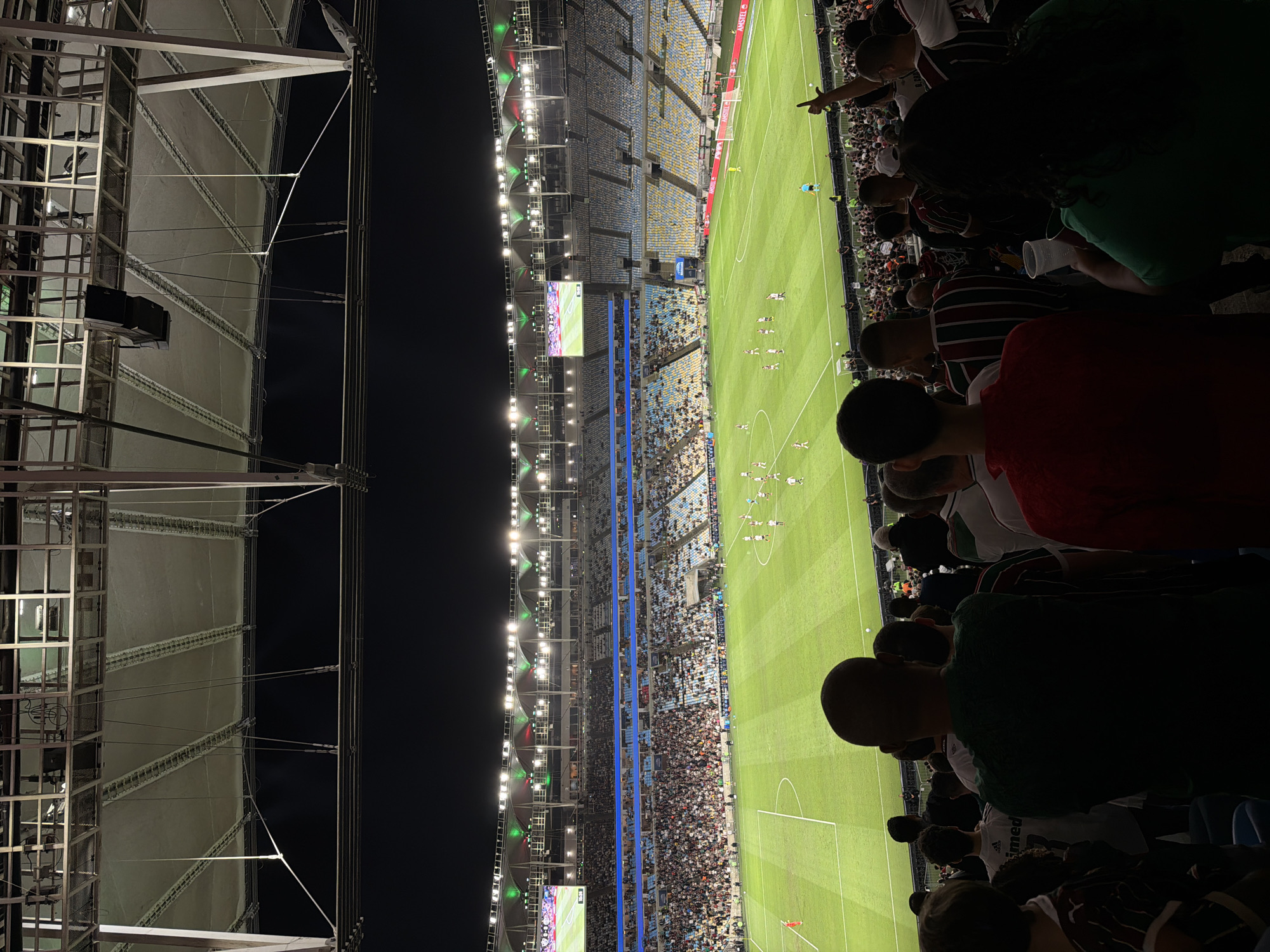 The image shows a soccer match at night in the Maracanã Stadium in Rio de Janeiro, Brazil. The stadium is brightly lit, and a large crowd of spectators fills the stands. The field is green and well-maintained, with players in red and white uniforms visible on the pitch. The image is taken from a high vantage point, looking down at the field and the surrounding stadium. The Maracanã Stadium is a famous landmark in Rio de Janeiro, known for its historical significance in the world of soccer.