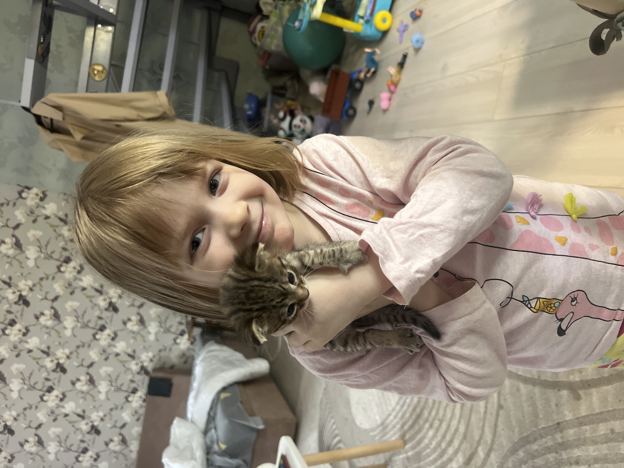 A young, light-skinned child with blonde hair is holding a tabby kitten. The child is smiling at the camera and is wearing a light pink, long-sleeved shirt with flamingo designs. The kitten is brown and black striped and appears to be a young animal. The background shows a room with floral patterned wallpaper on one wall, a set of stairs and a coat hanging on the stairs. Toys are scattered on the floor. The room appears to be a child's playroom or living space. It is difficult to confirm that the location is Chaikovsky, Russia based on the image alone. More context would be needed to confirm the location.