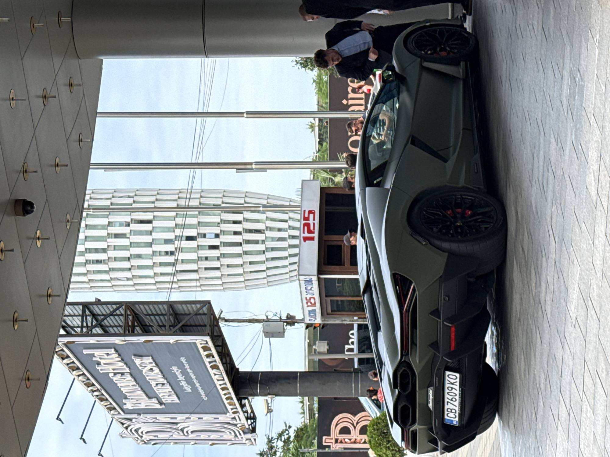 A matte green Lamborghini is parked on a patterned paved surface in front of a building with large glass windows and a modern, patterned tower in the background. A large, illuminated billboard for "Billionaire Hotel" is visible to the left. Several men are standing near the car. One man in a suit is bending down, appearing to adjust his trousers, while others stand around him. To the right of the car, a sign reads "125". The sky is overcast.