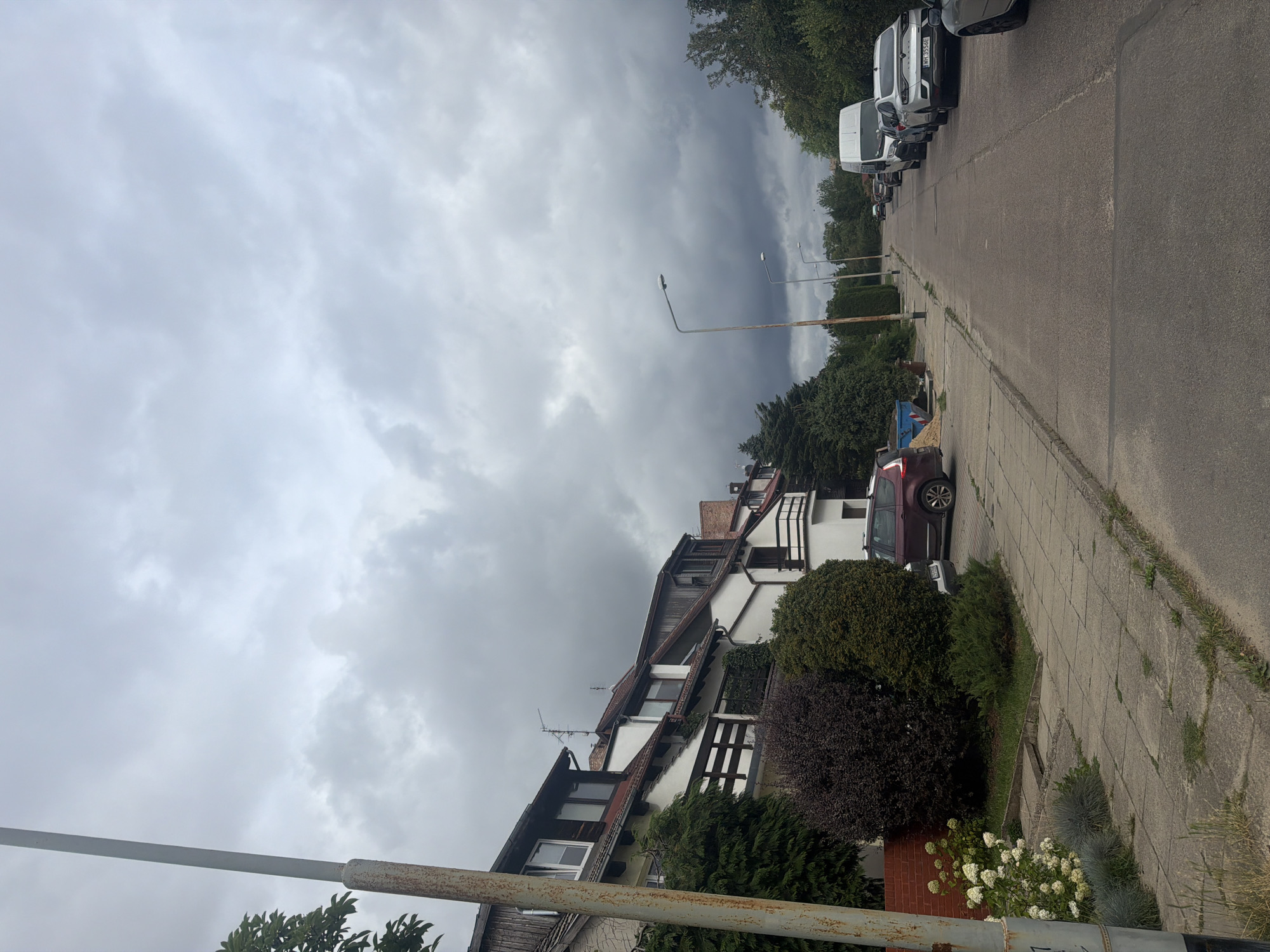 The image depicts a residential street in Szczecin, Poland, under a heavy, overcast sky. The street is lined with two-story townhouses on the left, featuring white facades, dark-tiled roofs, and wooden balconies. Lush green trees and dark purple bushes are planted in front of the houses. The sidewalk is paved with gray concrete tiles, and there is a paved road on the right side of the image. Several cars are parked along the road, including a white van, a silver sedan with Polish license plates, and a dark red SUV. Streetlights are visible along the road. The overall scene suggests a quiet, residential neighborhood on a cloudy day.