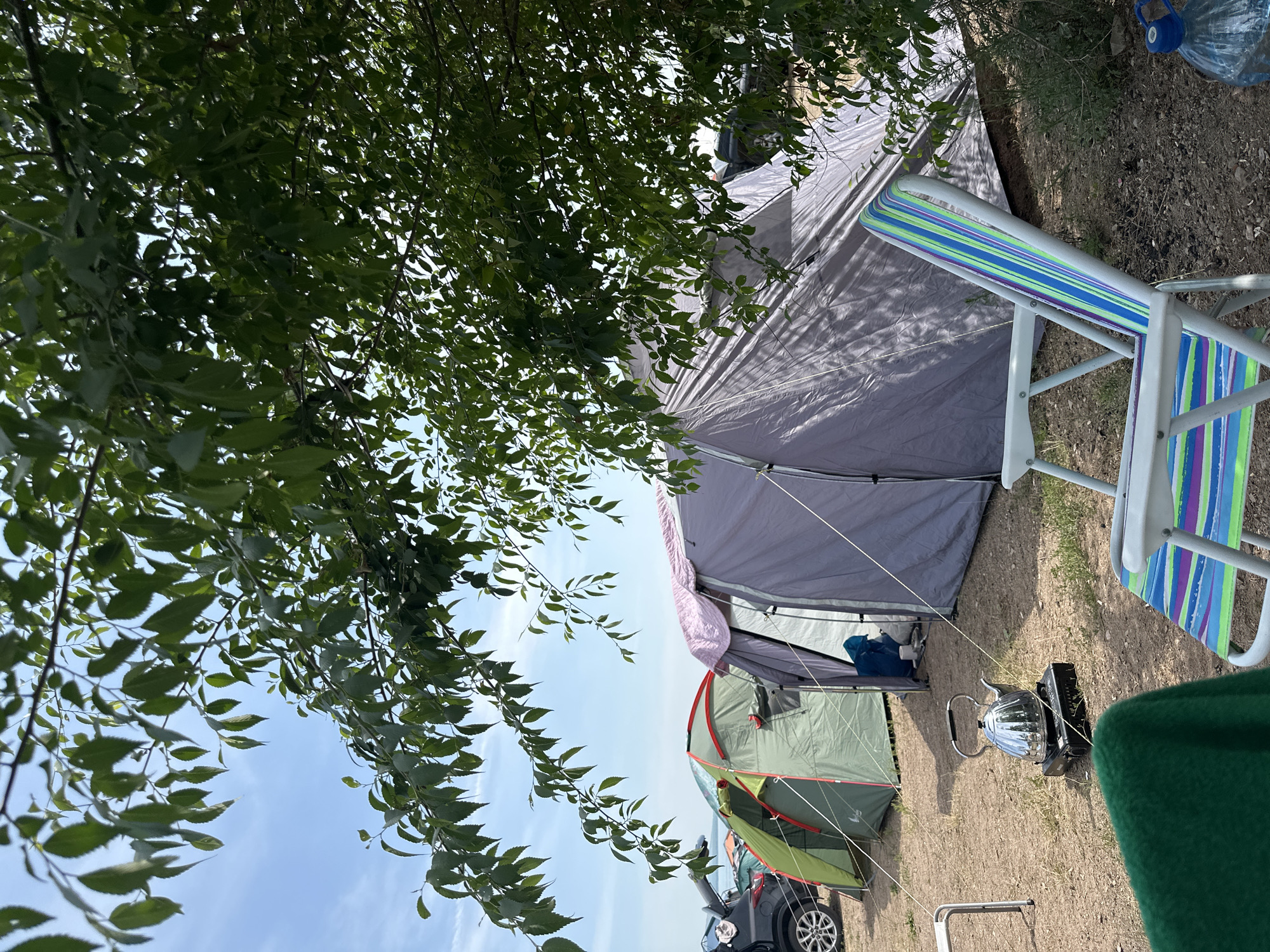 The image depicts an outdoor campsite situated near a body of water under a clear blue sky. The foreground features a colorful striped camping chair and a green blanket draped over another chair. Further back, a metallic kettle sits on a portable camping stove. Several tents are pitched on the sandy ground, including a large grey tent and a smaller green tent with a red trim. Behind the tents, the rear of a black car with its trunk open is visible. The campsite is partially shaded by the leaves of a tree, whose branches extend into the top of the frame. The setting appears to be a tranquil location, possibly a park or recreational area, in or near Kolodezny, Russia. There are no people visible in the image.