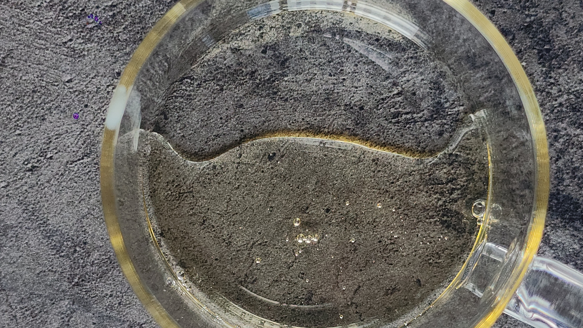 An overhead shot depicts a clear glass mug, partially filled with liquid, resting on a rough, gray surface. The mug has a gold-colored rim and handle. The liquid level in the mug is uneven. There are several small bubbles visible within the liquid. The gray surface appears to be textured and granular. There is no information available to suggest the image was taken in Moscow, Russia. There are no people present in the image.