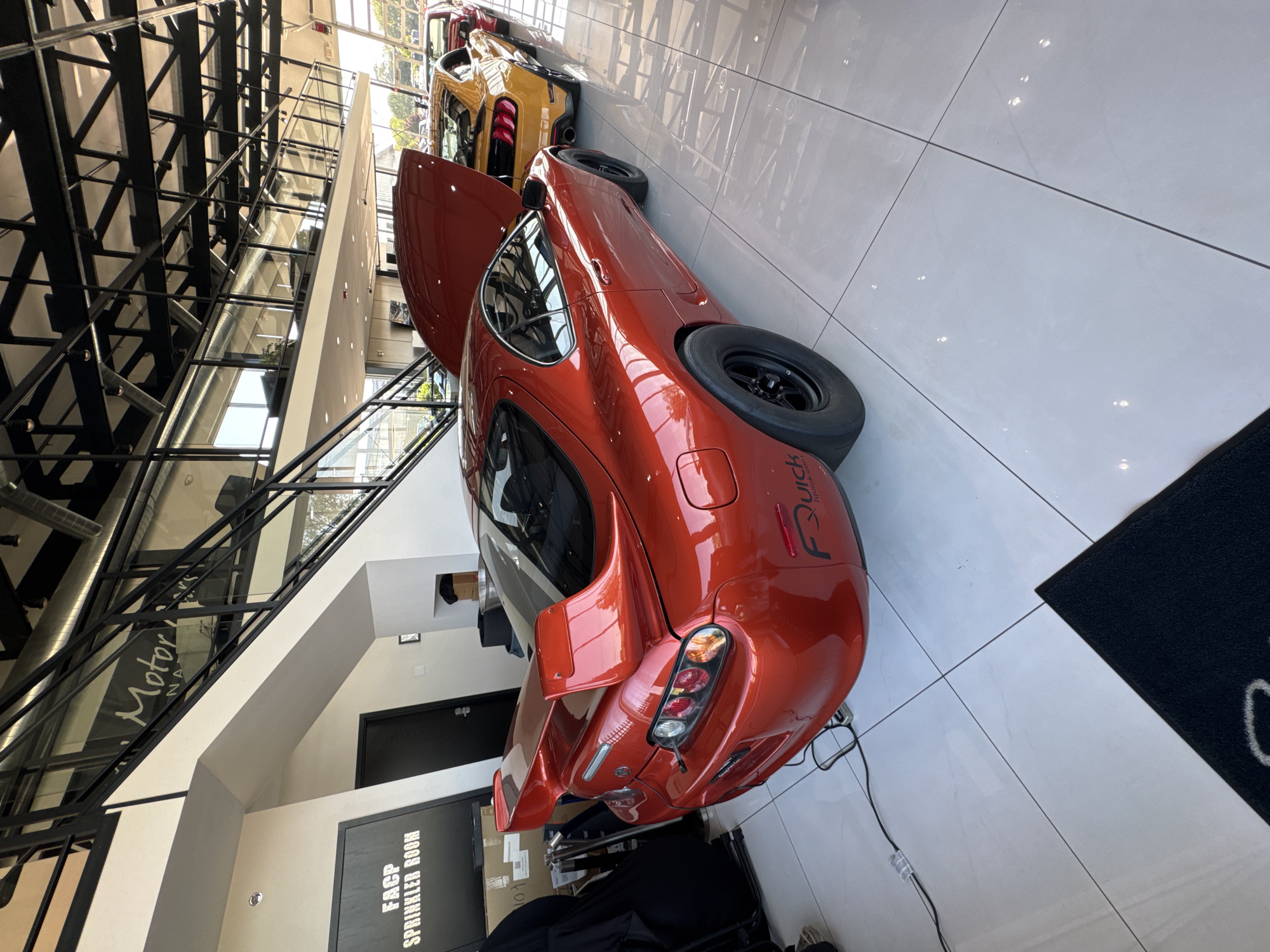 The image shows the interior of a building in Naperville, Illinois, United States of America, with an orange Toyota Supra and a yellow Mustang parked on a white tiled floor. The Supra's hood is open. A black mat is on the floor near the car. A sign reads "FACP Sprinkler Room". Above the cars is a metal structure.
