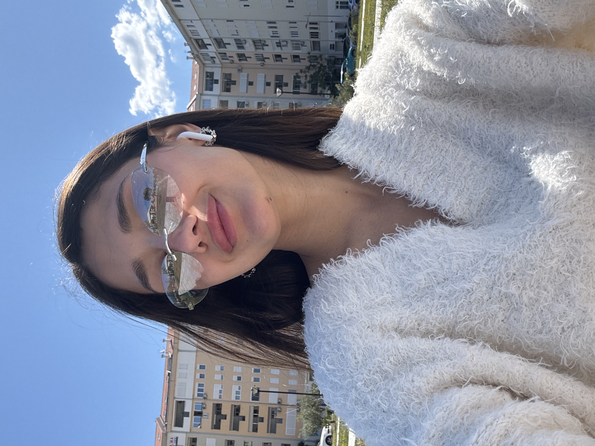 A young woman with long brown hair wearing frameless tinted sunglasses and a white fuzzy sweater is taking a selfie outdoors. She also has a white earbud in her left ear. Behind her are apartment buildings, blue sky, and some clouds. The buildings and clear sky suggest a location in Šušanj, Montenegro.