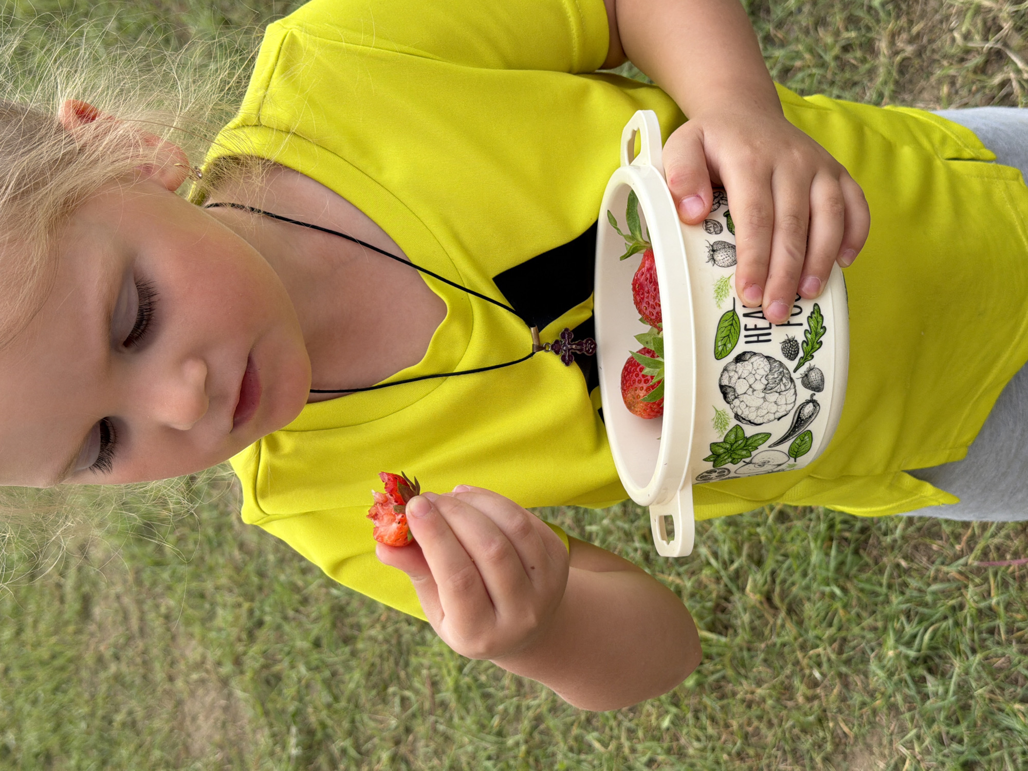 A young girl with blonde hair and eyelashes is holding a partially eaten strawberry in her right hand, and a bowl of strawberries in her left. She is wearing a bright yellow t-shirt and a necklace with a cross pendant. The background consists of green grass, suggesting an outdoor setting. The image was taken in the city of Chaikovsky, Russia.