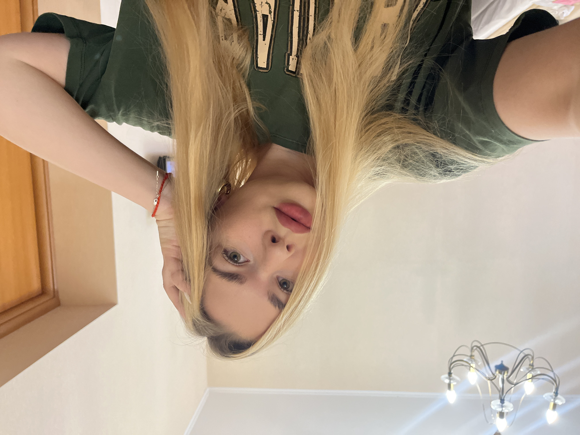 A young woman with long blonde hair and makeup is taking a selfie in an indoor setting. She is wearing a green t-shirt. She is touching her hair with her right hand. There is a chandelier visible in the background. The walls are a light color. A wooden door is visible on the right side of the image.