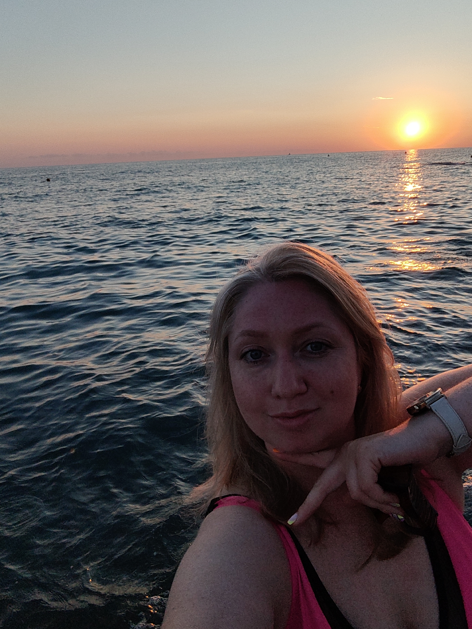A woman takes a selfie in the sea at sunset. The woman has blonde hair and is wearing a pink and black swimsuit. She is wearing a watch on her left wrist. The sea is calm with small waves. The sun is setting over the horizon, casting a golden glow on the water and the sky. The sky is a gradient of orange, pink, and blue. The photo appears to have been taken in Sochi, Russia, given the context of the image, which is likely a tourist destination with beaches and a sea.