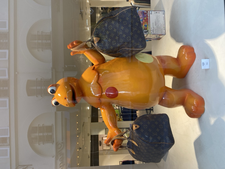 The scene displays a retail storefront window in Paris, France, featuring a large, glossy orange, bipedal, frog-like character statue. The statue, with prominent round eyes, a wide mouth, a red circular detail on its chest, and a lighter circular detail on its lower right side, holds one Louis Vuitton Monogram canvas "Speedy" style bag in each outstretched hand. The bags exhibit worn leather handles and trim. On the white display floor in front of the statue, a small rectangular sign is visible, bearing the text "CAIMAN" and "700€". Behind the statue's right leg, a white box with "SPACE CADES" printed on it is partially visible. The clear glass of the display window strongly reflects the exterior environment: a classic Parisian building facade with arched windows, decorative balconies, and white louvered shutters, alongside reflections of interior store elements such as clothing racks and overhead lighting. No people are visible within the immediate display or clearly in the reflections.