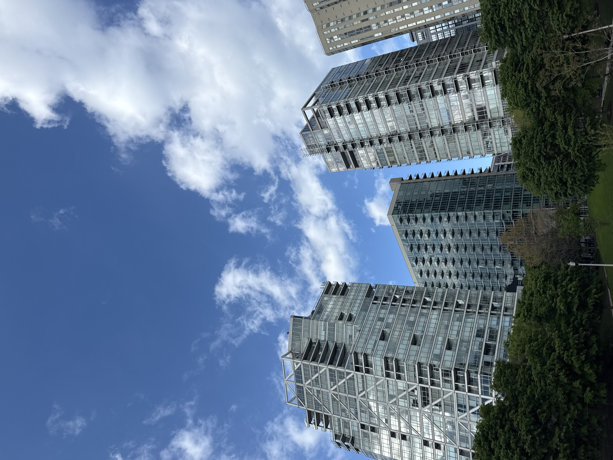 The image shows a low-angle view of several high-rise buildings against a bright blue sky dotted with puffy white clouds. The buildings appear to be modern architectural designs, primarily constructed of glass and metal. Some are residential apartment buildings. Green trees are visible along the side of the image, suggesting a park or green space. Based on the architectural style and general layout, this photo was taken near Lakeshore East in Chicago, USA. There are no people visible in the image.
