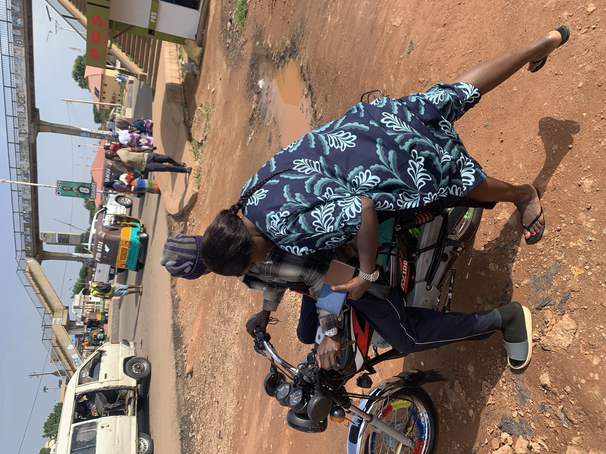 The image depicts a street scene in Gura Top, Nigeria, with a focus on a man and a woman on a motorcycle. The man, wearing a plaid shirt and dark pants, is seated on the motorcycle, holding the handlebars and looking forward. The woman, dressed in a dark blue and teal patterned outfit, is seated behind him, holding a blue wallet and a brown notebook. Her arm is extended, possibly indicating an action or interaction. The motorcycle, a black AX100 model, is parked on a reddish-brown dirt road.
In the background, other vehicles are visible, including a white van and a yellow and blue auto-rickshaw. Pedestrians are present, including a man in a green shirt and dark pants, talking on his phone, and a person wearing a yellow vest and blue shorts. A pedestrian bridge crosses over the road in the distance, adorned with graffiti. The environment is characterized by dusty roads, scattered debris, and a puddle of muddy water on the ground. The overall impression is one of a bustling, informal urban setting.