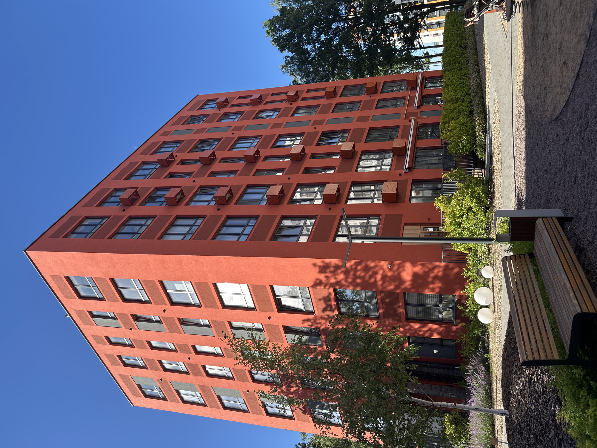 The media file depicts a modern, tall, salmon-colored residential building with numerous windows and square air conditioning units protruding from its facade. The building's architectural style is geometric and repetitive. To the right of the building, there is a paved walkway bordered by gravel and lush green landscaping, including shrubs, trees, and flowering plants. A wooden bench is visible in the foreground, adjacent to the walkway. The scene is illuminated by bright sunlight, suggesting it is daytime. The sky is a clear, vibrant blue, indicating good weather. The overall impression is one of contemporary urban living with integrated green spaces. The context provided places this scene in Kaluga, Russia. There is no visible text in the image.