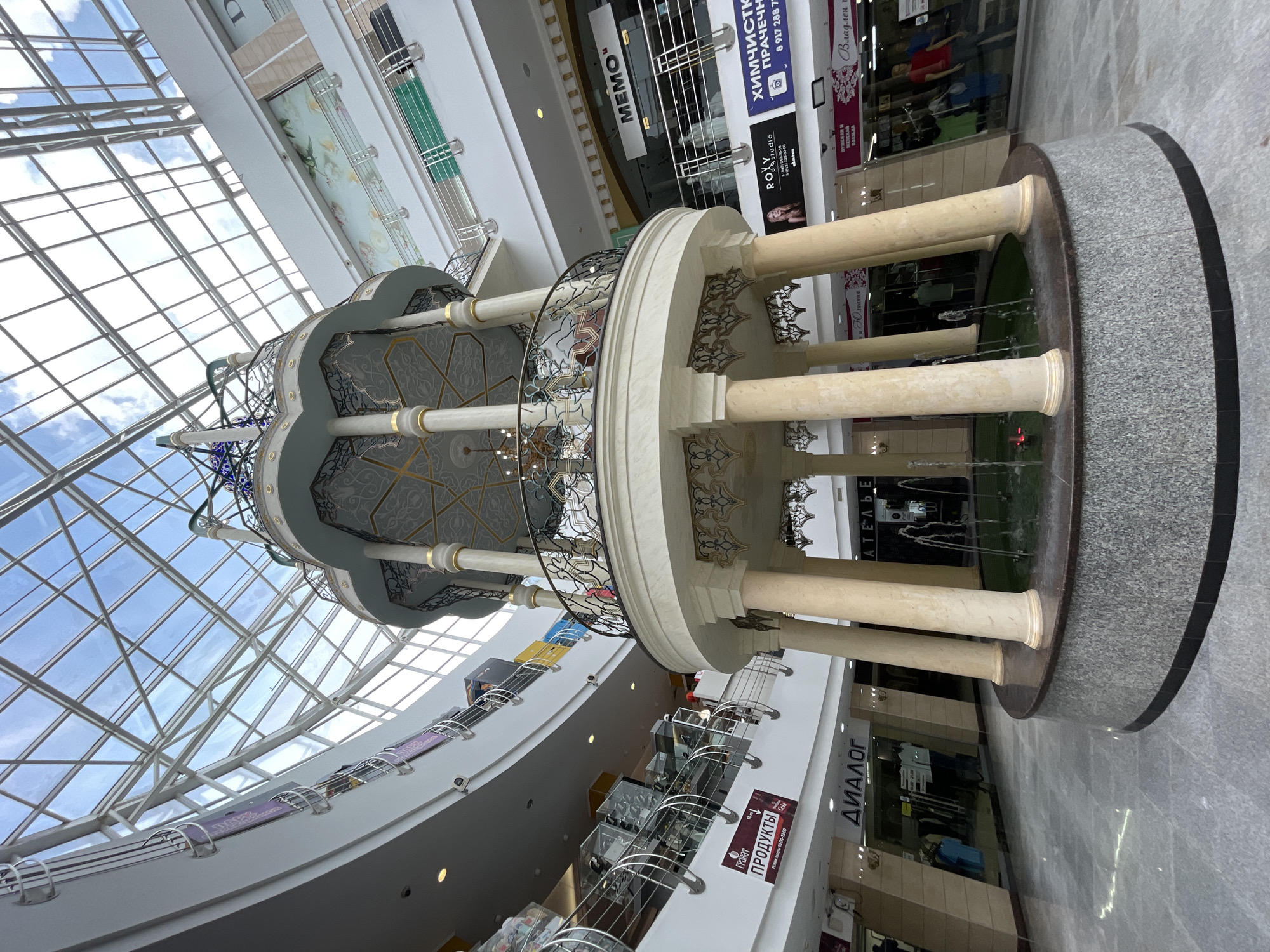 The image shows the interior of a modern shopping mall, likely in Kazan, Russia, with a focus on a central decorative fountain structure. The structure is multi-tiered, featuring a circular base with fountains, supported by large, fluted columns. Above the columns, there is a balcony with ornate railings and a decorative roof. The mall levels surround this central feature, with shops visible on each level. The ceiling of the mall is a large glass and metal skylight, allowing natural light to illuminate the space. Shops visible include "Диалог", "Продукты", "MEMO", "ROXY studio", and "Химчистка". The floor is tiled with a polished grey stone. The environment appears clean and well-lit, with no people prominently featured in the foreground.
