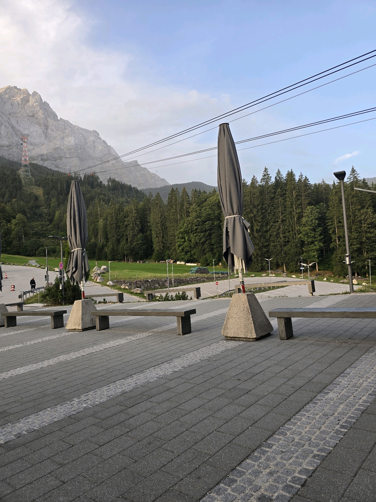 The image depicts an outdoor scene in Grainau, Germany, likely at a cable car station for the Zugspitze. In the foreground, a paved area is visible, composed of dark gray cobblestones with lighter gray striped accents. Several wooden benches with dark gray metal legs are arranged along the paved area. Two large, closed gray umbrellas stand upright, each mounted on a pyramidal concrete base.
In the midground, a parking lot is visible with white lane markings. A person in a dark jacket and dark pants is walking away from the camera on the edge of the parking lot. Adjacent to the parking lot is a well-maintained green lawn bordered by a low stone wall.
The background features a dense forest of tall, dark green pine trees covering the slopes of a large, majestic mountain range. The mountain peaks are rocky and light gray, with some snow or mist visible near the summit. A red and white tower, likely part of a cable car infrastructure, is situated on the mountainside. Multiple parallel dark cables, indicative of a cable car system, stretch across the sky from left to right. The sky is a pale blue with scattered white clouds. The overall atmosphere suggests a tranquil, natural setting with man-made amenities.