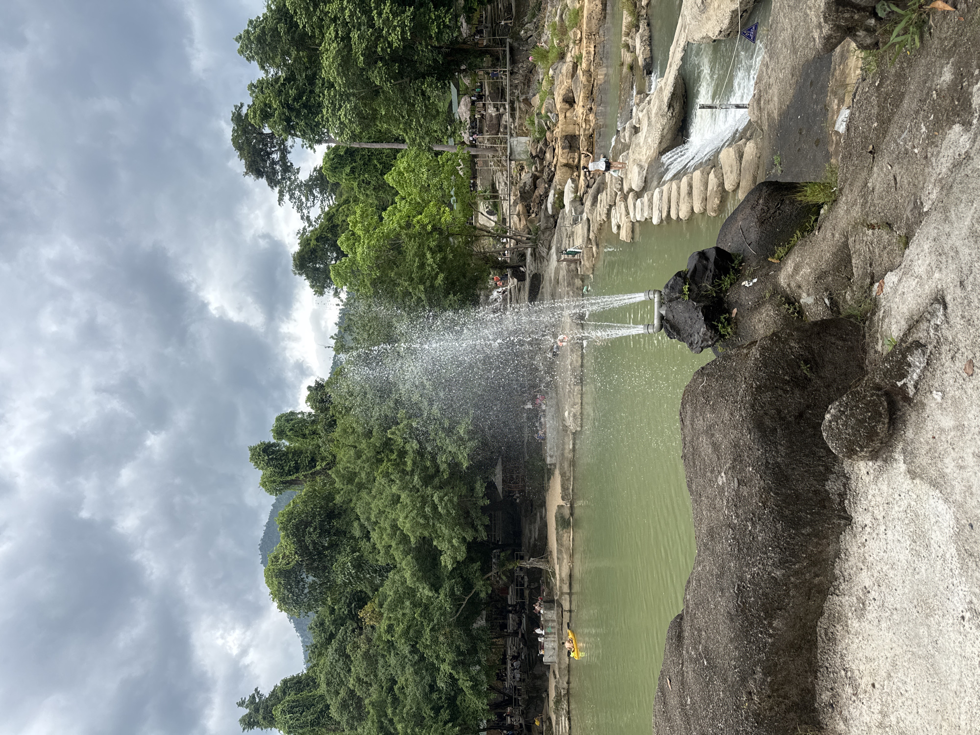 The image depicts a scenic view of a natural river flowing through a lush green environment in Khanh Vinh, Vietnam. The river is a prominent feature, with calm, light green water occupying a large portion of the foreground and mid-ground. In the mid-ground, a water fountain sprays water upwards, adding a dynamic element to the scene.
A path made of stepping stones crosses the river, connecting the two banks. On the far bank, there is a sandy area with numerous people gathered, some sitting and others walking. Several thatched-roof structures, possibly shelters or small shops, are visible in this area, suggesting a recreational or tourist spot. A person is seen kayaking in the river on the left side of the image.
The surrounding environment is dominated by dense green trees and vegetation, with a hilly or mountainous backdrop. The sky is overcast with heavy, grey clouds, indicating a cloudy day. Large rocks are scattered in the foreground and along the riverbanks, some covered with moss. The overall impression is one of a natural, somewhat developed recreational area within a verdant landscape.