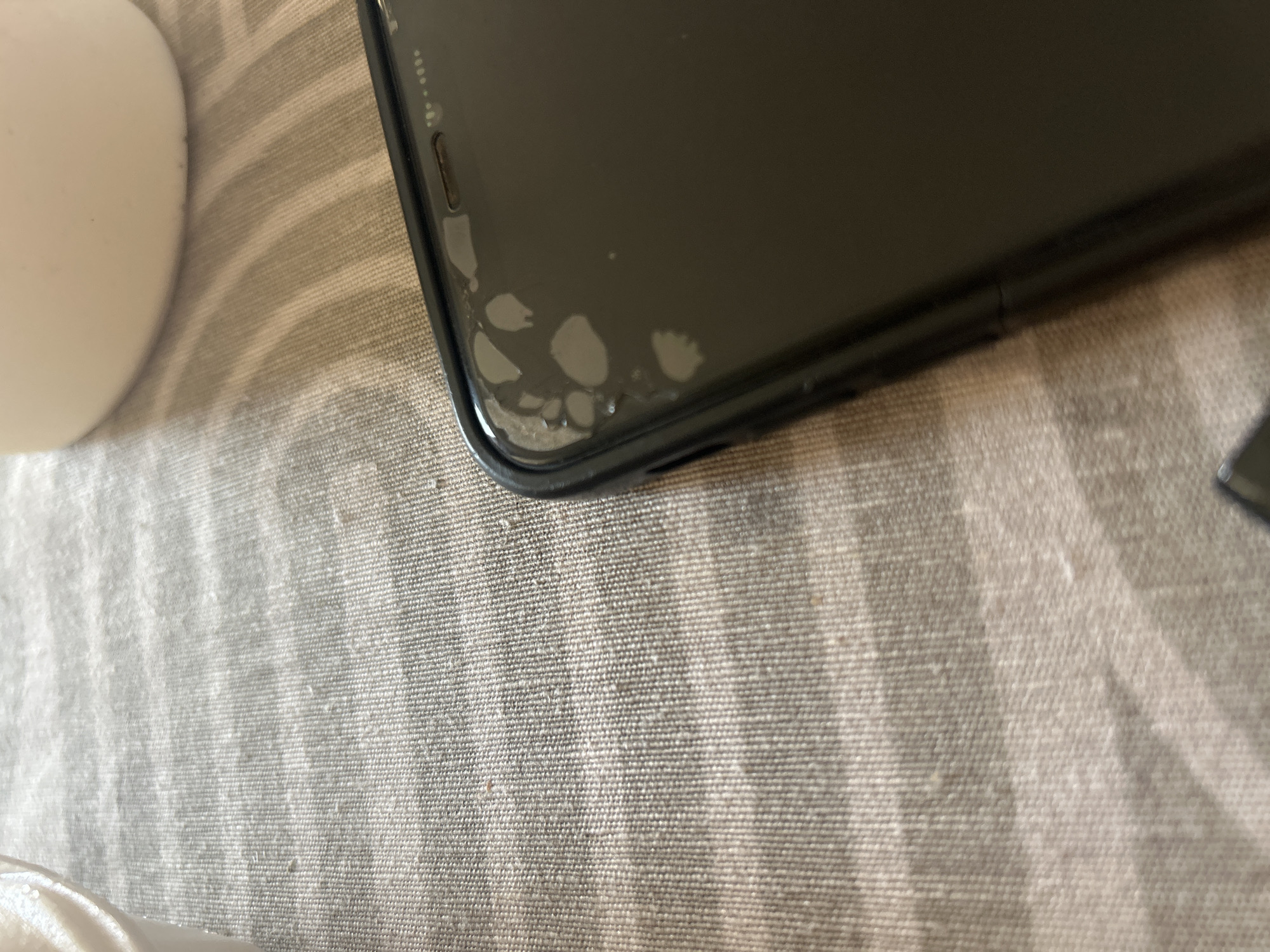 The image shows a close-up of a smartphone screen with several air bubbles or damage marks on the top left corner, against a textured, striped fabric surface. The background includes a portion of a white, curved object, possibly a cup or container. The scene is indoors, likely on a table or countertop. There is no indication of location in Plovdiv, Bulgaria.