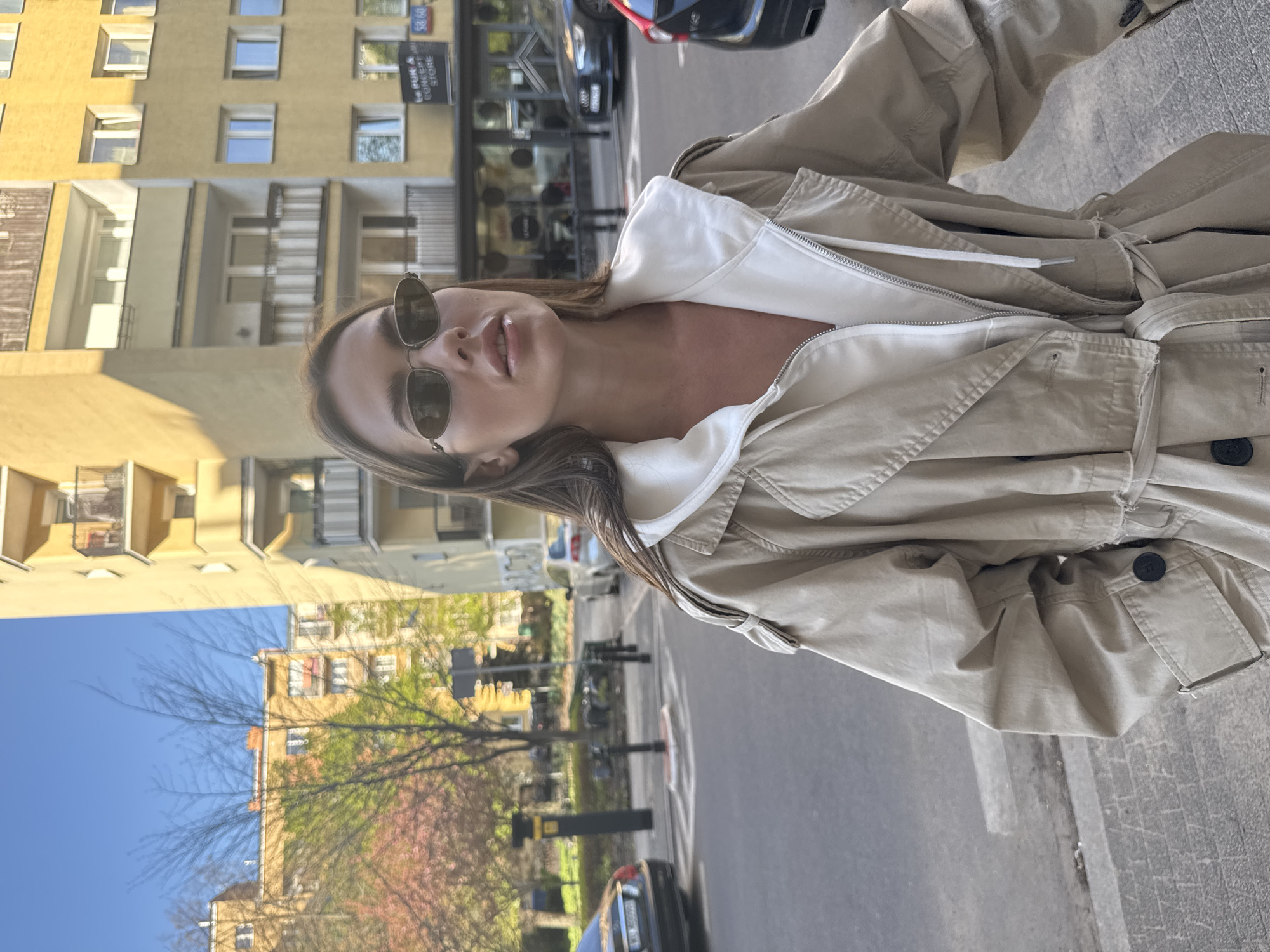 A bright, sunlit, medium-shot image captures a woman on an urban street. She has long brown hair and is wearing dark sunglasses, a beige or khaki trench coat layered over a white hooded top. Her face is tilted slightly upwards, with her lips subtly parted.
The background depicts a street scene in what is identified as Warsaw, Poland. To her right, a tall, light yellow building with multiple windows and balconies is prominent. The street extends into the distance, lined with parked cars and trees. Some trees have bare branches, while others display delicate pink blossoms, indicative of spring. The sky above is clear and bright blue.
A dark sign on a building in the background is legible, displaying "LE BUK A CONCEPT STORE" and the numbers "58/60". The strong, natural lighting suggests it is daytime, under clear weather conditions. The ground features asphalt pavement and concrete sidewalks. The overall aesthetic is casual and reflects street style.