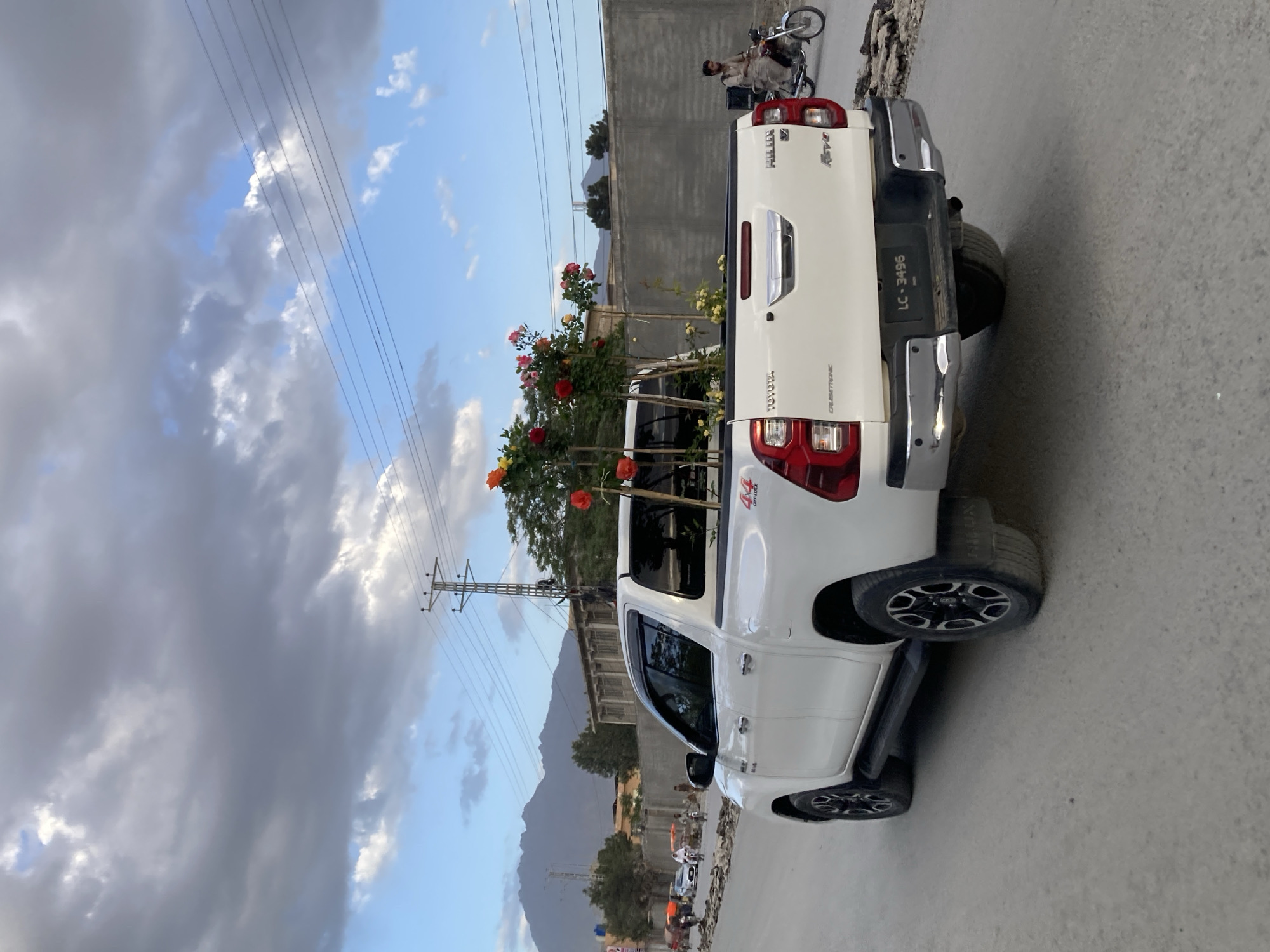 A white Toyota Hilux Revo pickup truck is parked on a paved road in Quetta City Tehsil, Pakistan. The truck bed is carrying a shrub with red and orange flowers. A motorcycle rider is visible behind the truck, and a building is in the background. Mountains and power lines are visible in the distance under a partly cloudy sky. The license plate of the truck reads "LC-3496".