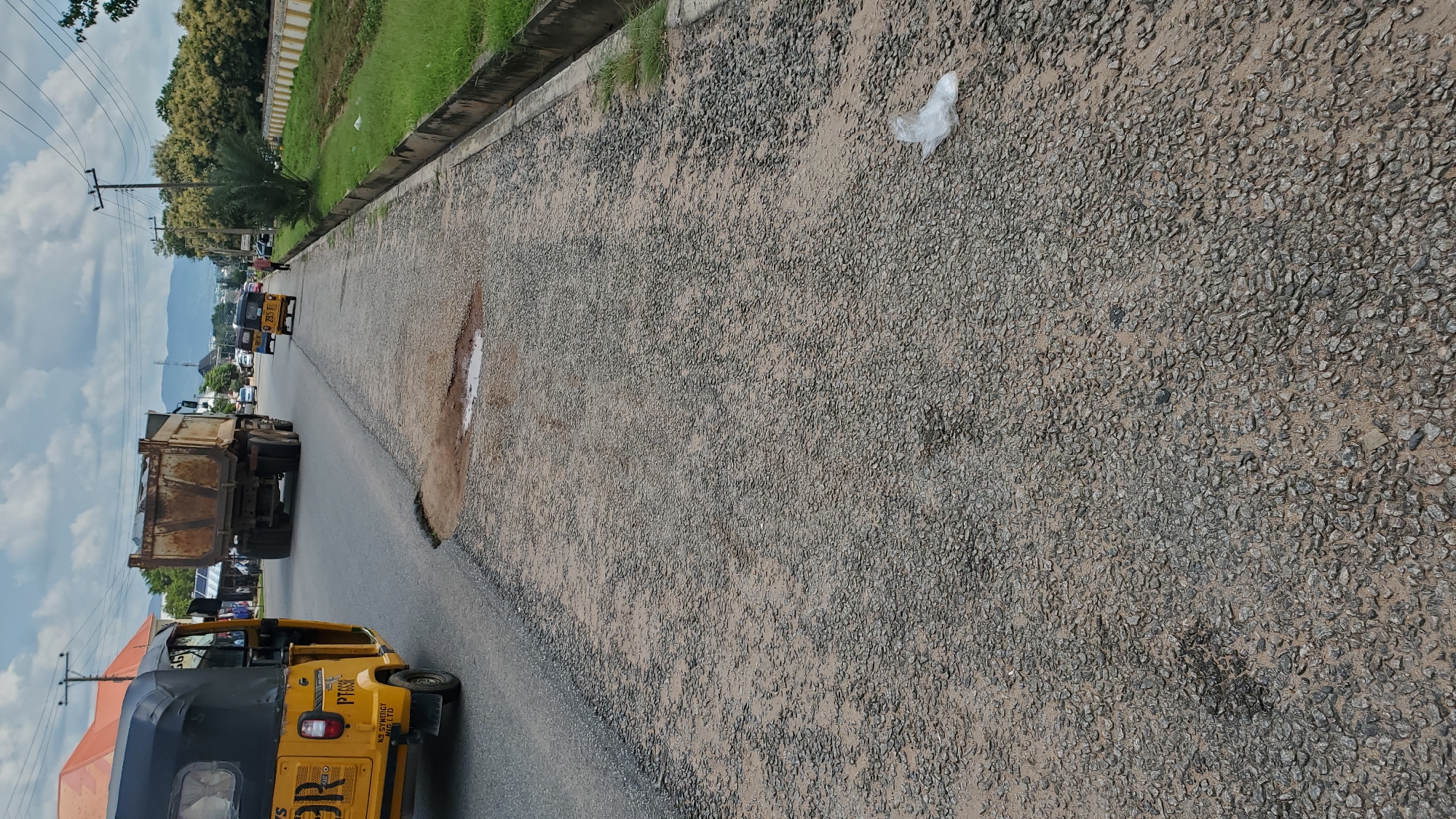The image depicts a street scene in Jalingo, Nigeria. A yellow motorized tricycle, known as a "Keke Napep," is prominently featured on the left side of the road, with its registration plate visible. Further down the road, a large dump truck is driving away from the camera. Several other vehicles, including another Keke Napep, are also on the road in the distance.
The road itself is in a state of disrepair, with a large pothole visible in the foreground, containing some standing water. The surface of the road is uneven and appears to be made of gravel and asphalt. To the right of the road, a grassy area is bordered by a concrete drainage channel. A white plastic bag is lying on the road surface near the right edge of the image. In the background, trees line the roadside, and hills or mountains are visible in the distance under a cloudy sky. Buildings and utility poles are also present along the street.