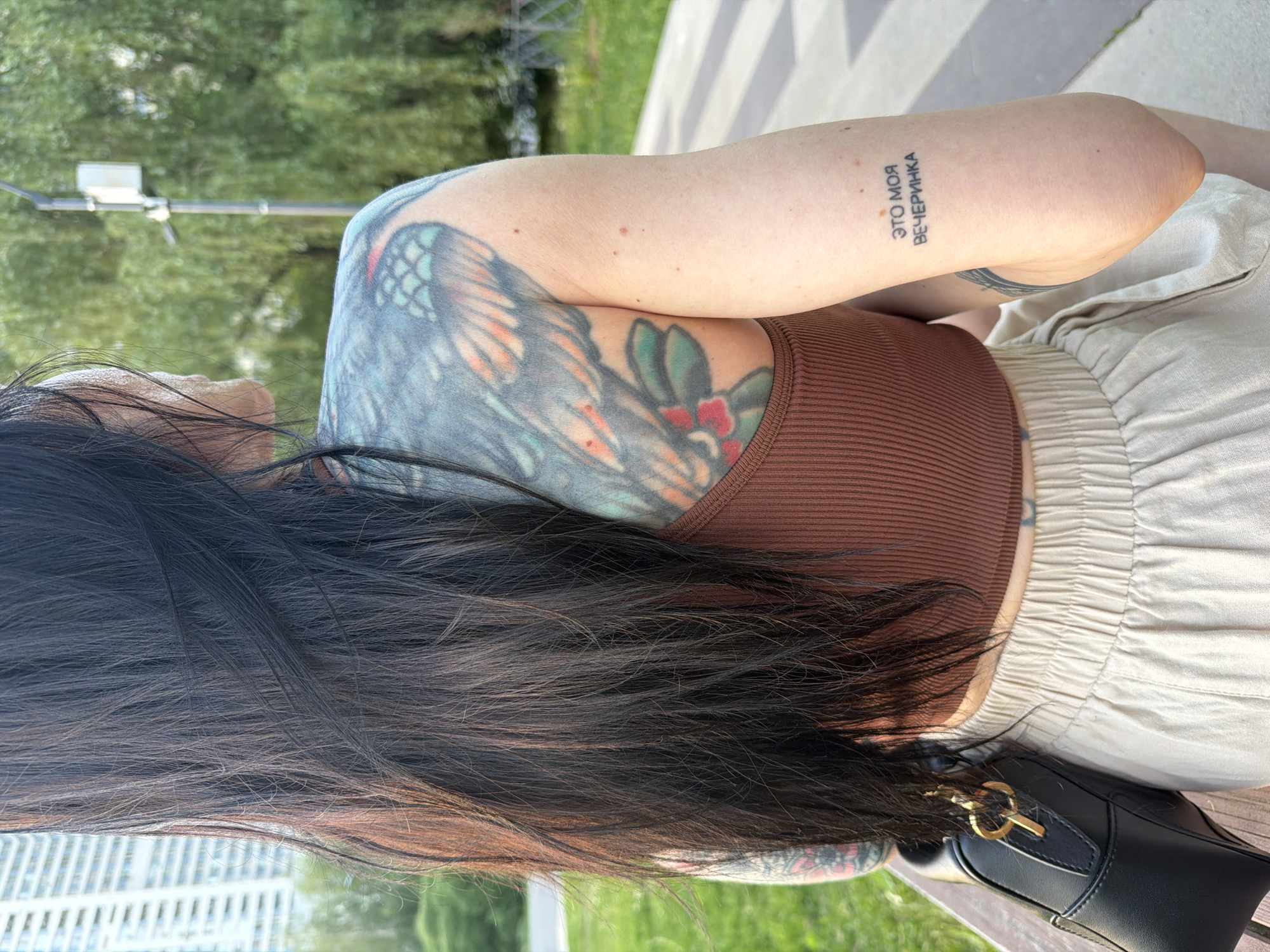 The image captures a woman sitting in a Moscow, Russia park. She has long, dark hair and wears a brown top and light-colored pants. A black purse rests beside her. Prominent tattoos are visible on her arm and shoulder. One arm has a tattoo in the Russian language reading "ЭТО МОЯ ВЕЧЕРИНКА," which translates to "This is my party." The location appears to be a well-maintained park with green grass and trees. A lamppost is visible in the background.