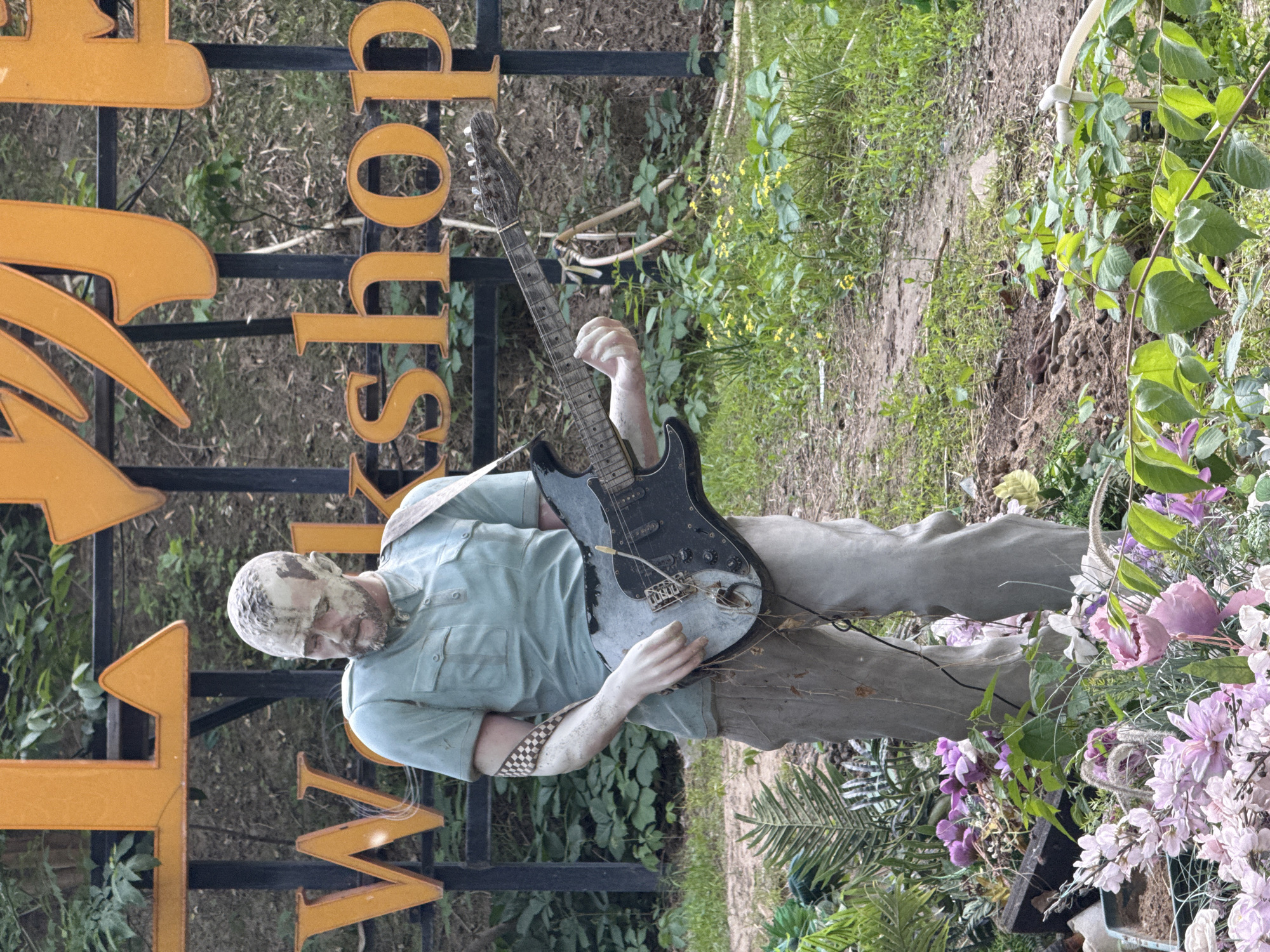 In an outdoor setting in Chaoyang District, China, a statue of a man is positioned playing an electric guitar. The statue is dressed in a short-sleeved shirt and pants, with a guitar strap visible. The statue stands adjacent to a sign with orange letters spelling "Workshop". The statue is situated within a garden environment featuring flowers and other greenery.