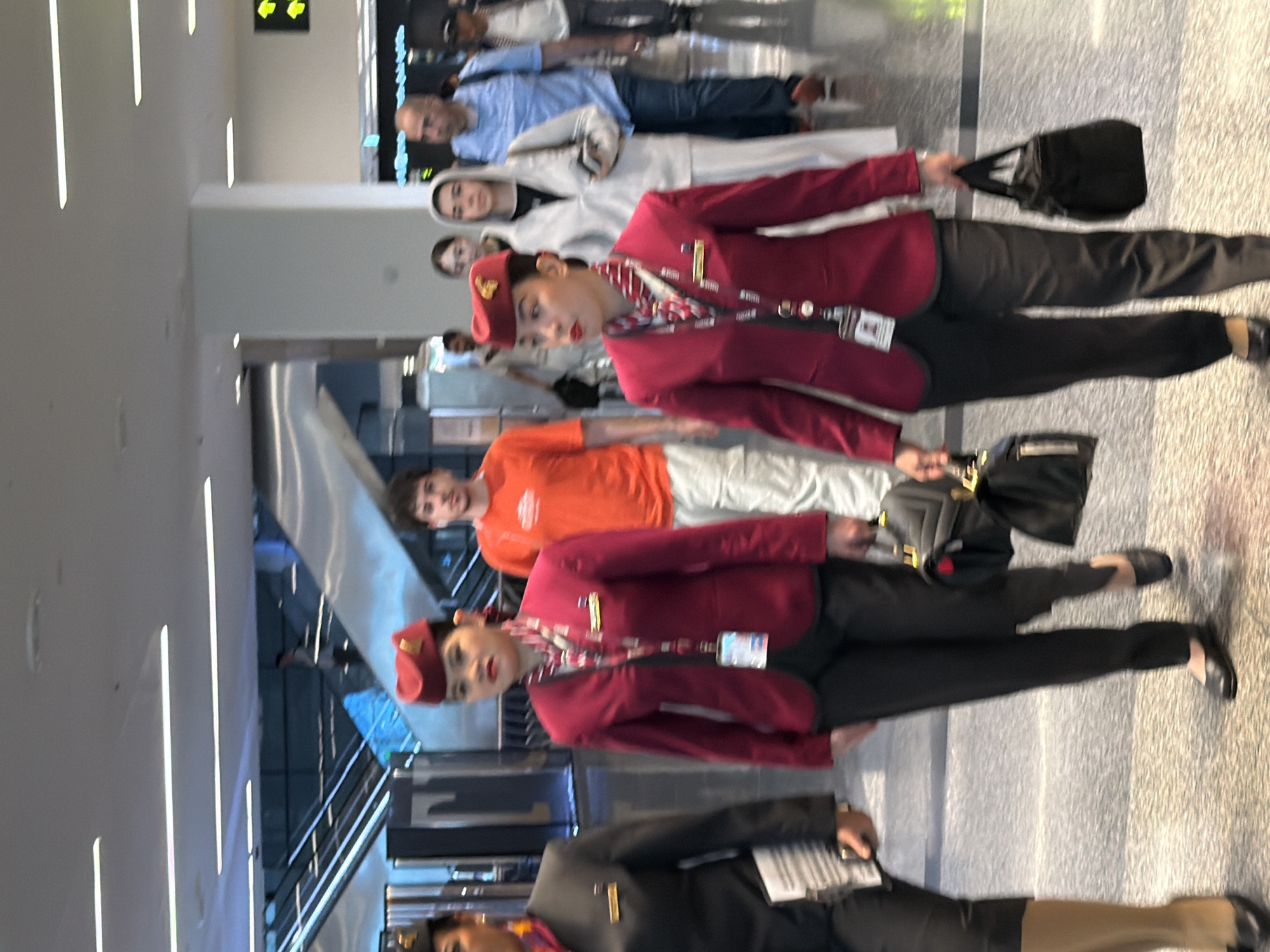 The image shows a group of people walking in an airport terminal. In the foreground, two women in maroon airline uniforms with red hats and scarves are walking side-by-side. The woman on the right is looking towards the left, while the woman on the left is looking forward. Both are carrying black handbags. Behind them, a man in an orange t-shirt is walking, followed by other travelers. The environment appears to be a modern airport with escalators and bright lighting. The scene is likely taking place in Hamad International Airport in Doha, Qatar, given the uniforms are similar to those worn by Qatar Airways staff.