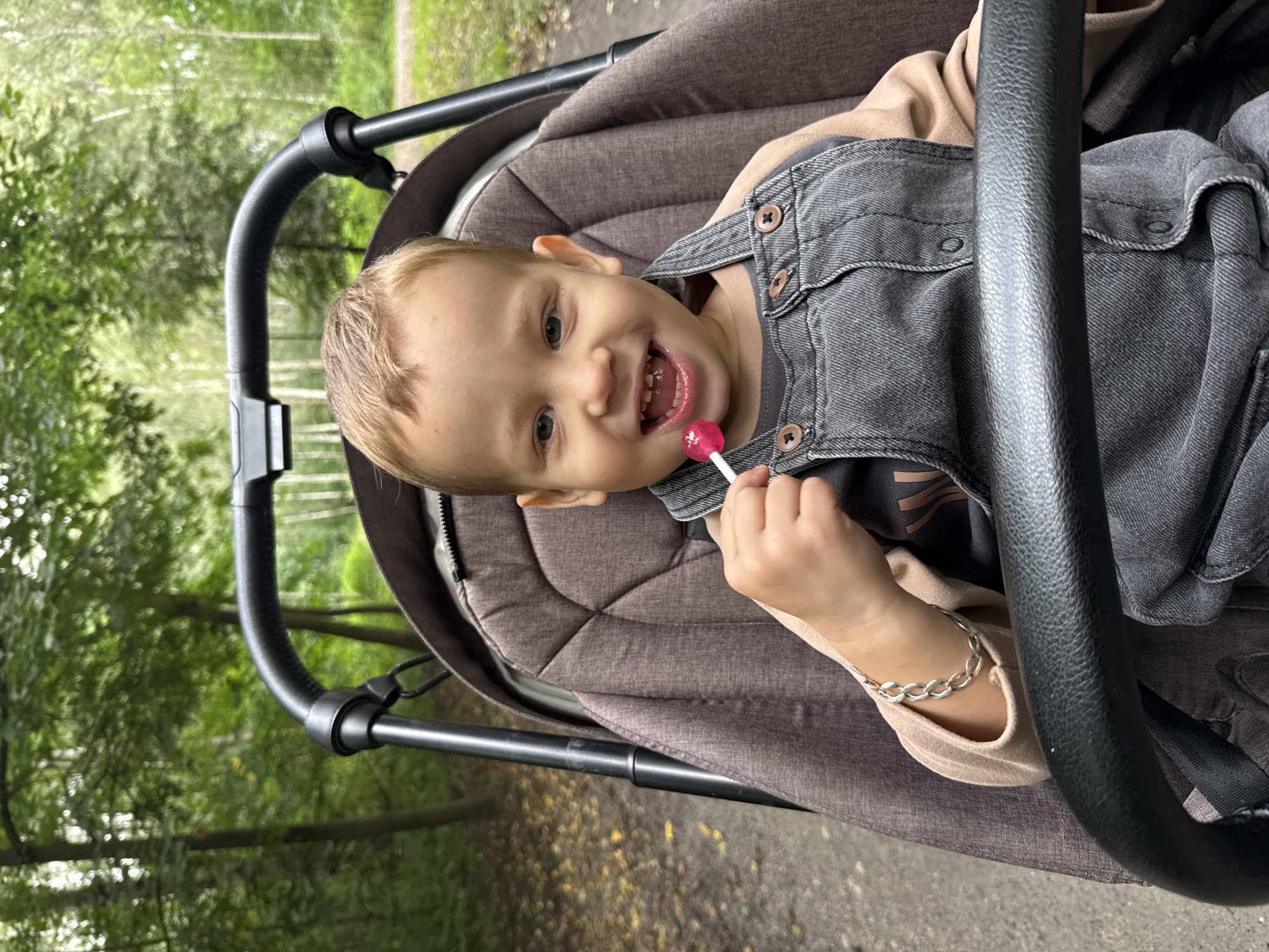 A young Caucasian child with blonde hair and blue eyes sits in a stroller, holding a pink lollipop to their mouth. The child is wearing a light brown long-sleeved shirt and dark gray denim overalls. They have a silver chain bracelet on their left wrist. The stroller is brown with a black handlebar. The background is a wooded area with trees and fallen leaves on the ground. The location is identified as Rusanovo, Russia.