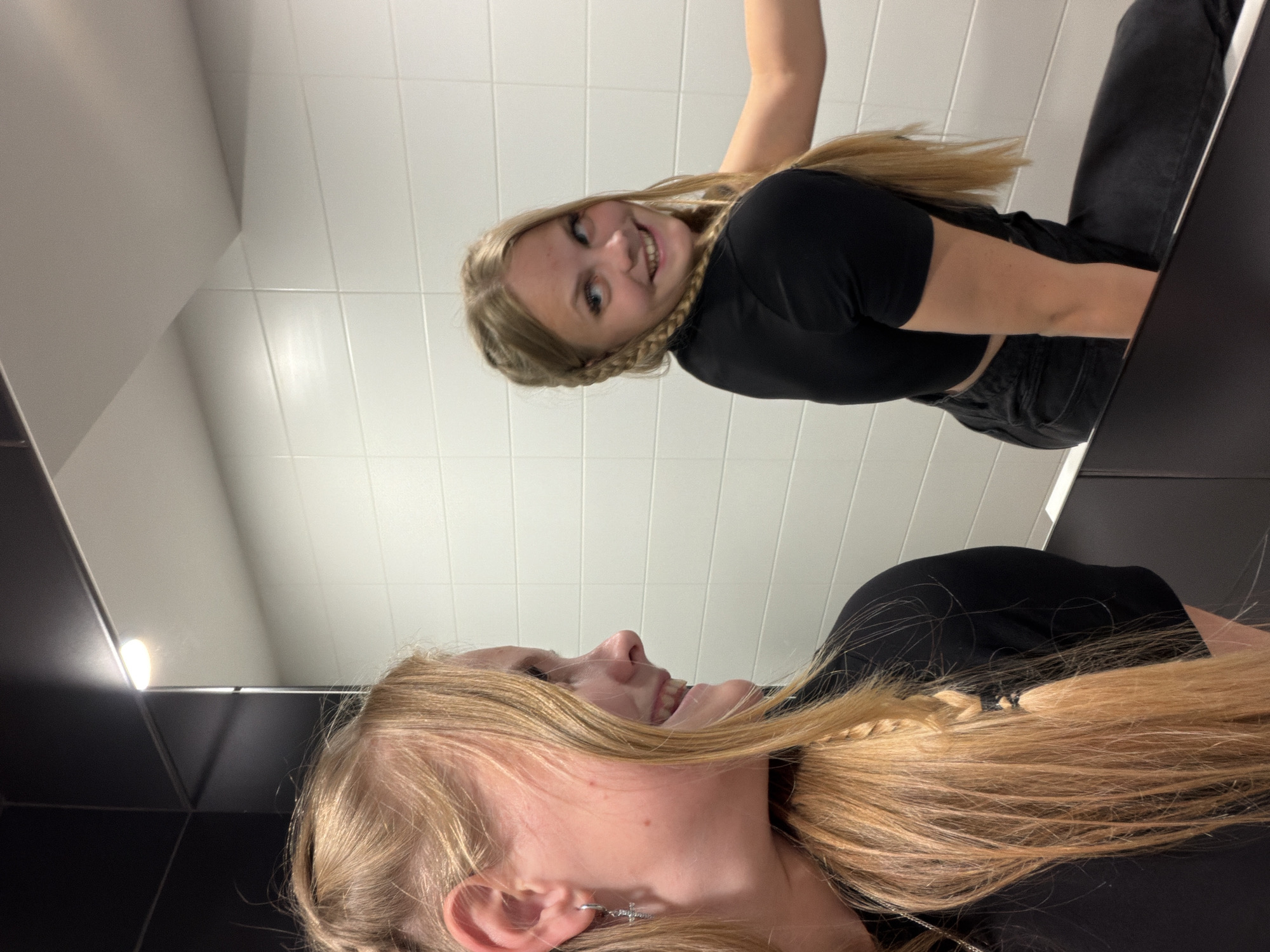 This media file shows two young women posing for a selfie in a brightly lit restroom. The setting appears to be a modern public restroom with white tiled walls and black fixtures, possibly in a mall or public building. One woman is in the foreground, looking sideways and smiling, with her blonde hair flowing around her face. She is wearing a black top and jeans, and a silver cross earring is visible. The second woman is in the background, also with blonde hair styled in braids, looking directly at the camera with a playful expression and a wide smile. She is wearing a black top and dark jeans. The reflection in the mirror suggests the photo is taken from a low angle, looking upwards. The lighting is bright and even, typical of interior lighting, and there are no indications of the time of day or weather. The overall mood is cheerful and informal, typical of a casual selfie. There is no visible text in the image.