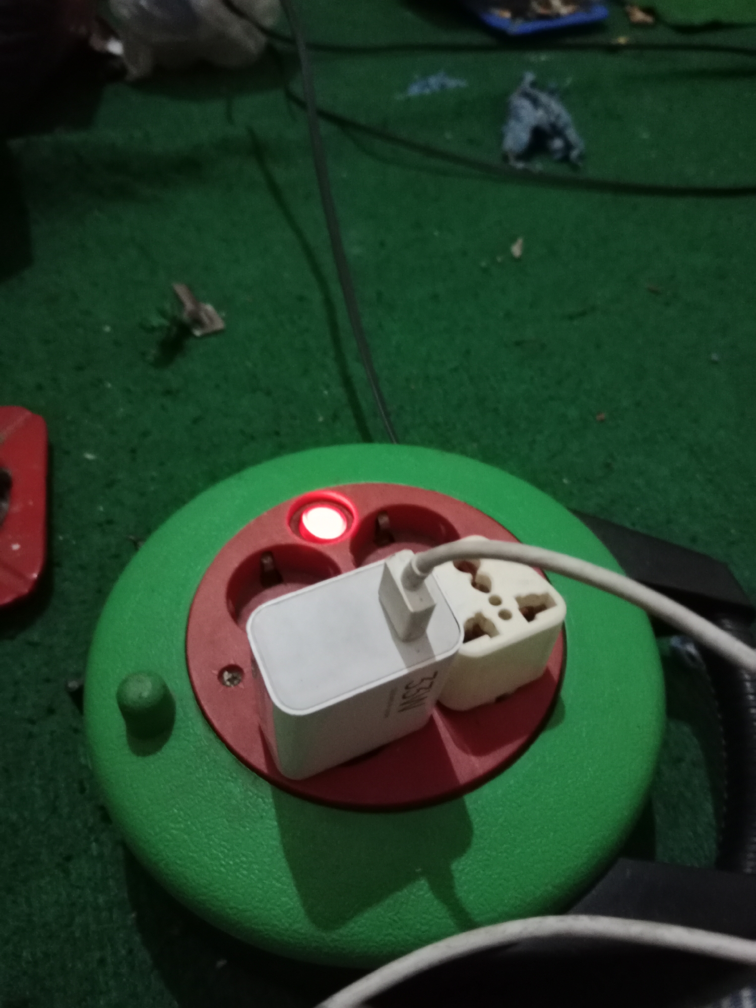 A circular green power strip with red accents is placed on a green, textured surface, possibly carpet or astroturf. The power strip has three visible sockets, one of which is illuminated by a red indicator light. A white power adapter, labeled with "33W," is plugged into one of the sockets, and a white cable extends from it. In the background, out of focus, are various objects, including what appears to be a blue tray, fabric scraps, and other indistinct items, consistent with a cluttered indoor environment. The overall lighting is dim, with a warm glow emanating from the illuminated socket. The scene is consistent with a domestic setting in Ringinrejo, Indonesia, where electrical accessories are commonly used.