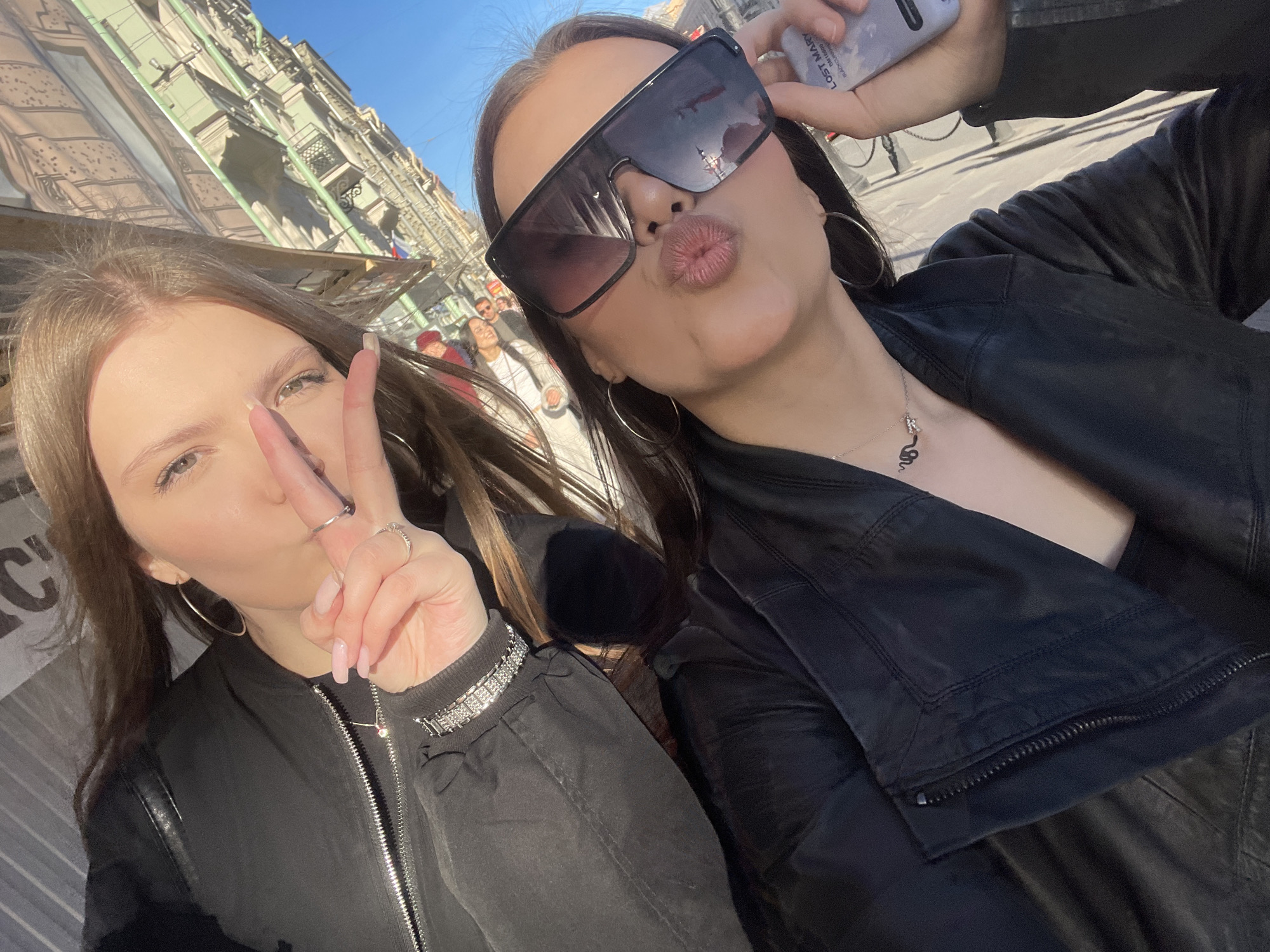 This media file is a bright, sunlit outdoor selfie featuring two young women, captured at a tilted angle against an urban backdrop. The clear blue sky and strong shadows suggest it is daytime.
The woman on the left has long, light brown hair and light-colored eyes. She wears a black bomber-style jacket with a silver zipper and a silver-toned bracelet on her left wrist. Her fingers are adorned with rings, and she displays light pink manicured nails. She is holding up a peace sign with her right hand, partially obscuring her face, while forming a subtle pout with her lips. She also wears silver hoop earrings.
The woman on the right has long, dark hair and is wearing large, dark square-framed sunglasses, which reflect the surrounding buildings. She is dressed in a black leather-look jacket and accessorizes with a silver necklace featuring a distinctive, curled pendant, resembling a stylized 'S' or snake. Her lips are puckered in a prominent kissy face. She is holding a light-colored smartphone in her left hand, likely taking the selfie. The back of the phone clearly shows the brand name "LOST MARY" in black lettering, accompanied by smaller text that reads "THE ONE AND ONLY" and "PUFF". She also wears gold hoop earrings.
The background depicts a street scene lined with tall, ornate historic buildings featuring balconies, characteristic of a European city like Saint Petersburg. Several other people are visible in the distant, out-of-focus background, some wearing hats. The overall atmosphere conveyed is casual and playful.