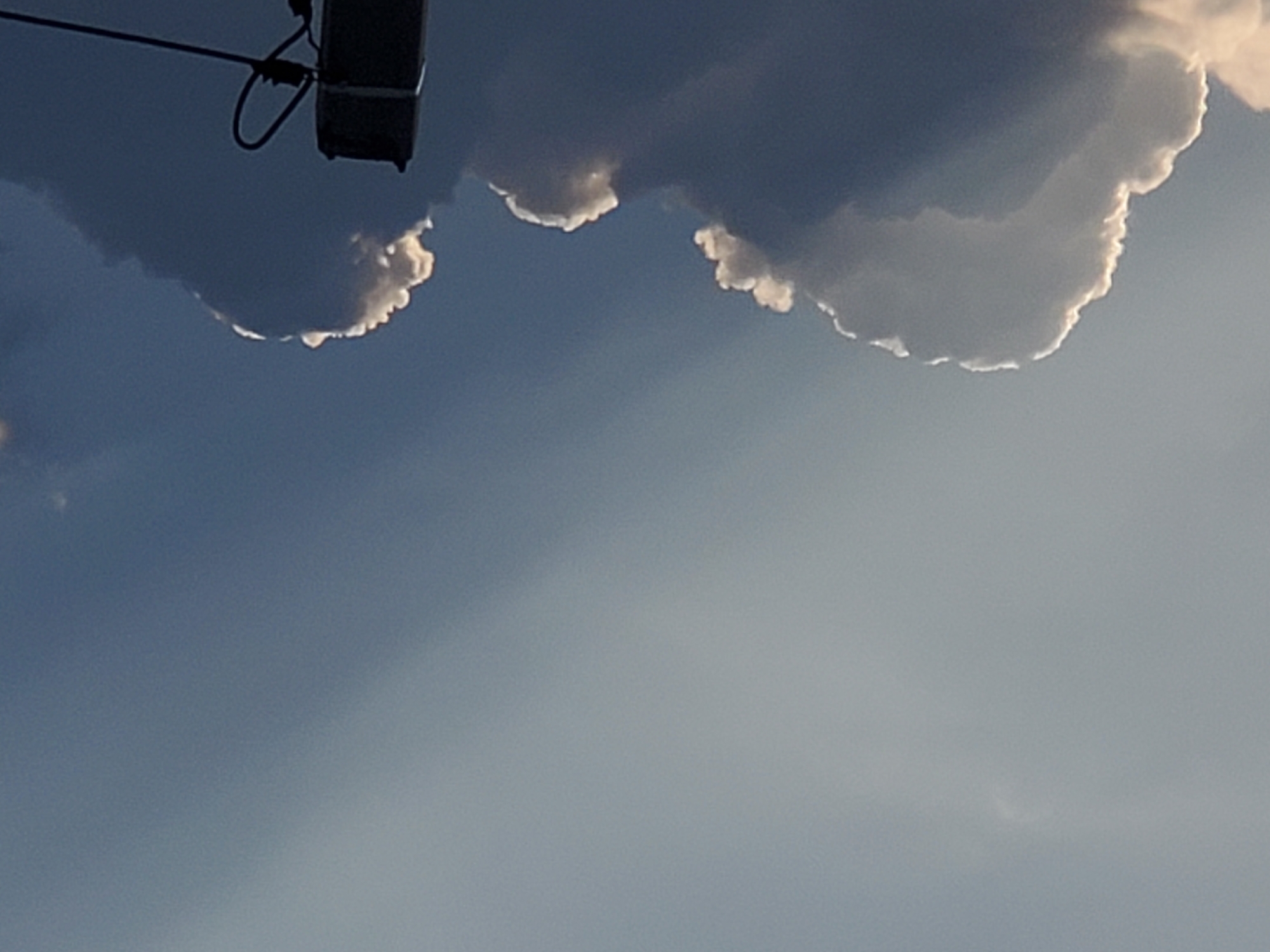 The image shows a cloudy sky with dramatic lighting effects. A beam of sunlight cuts through the darker clouds, illuminating some of them with a bright, white glow. The clouds themselves are large and volumetric, with some appearing soft and wispy while others are more dense and shadowed. In the lower right corner of the image, a dark, metallic pole with wires is visible, suggesting the presence of electrical infrastructure. The overall impression is one of a dynamic and atmospheric sky, possibly captured during a storm or at sunrise/sunset. There are no people or specific landmarks that would indicate a precise location within Konstantinovka, Moldova.
