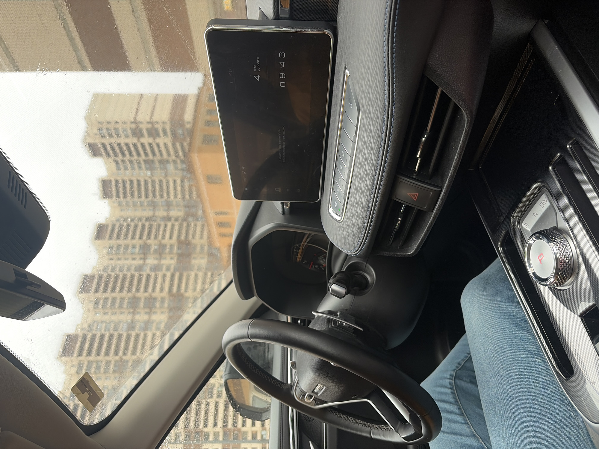 The image shows the interior of a car, specifically the driver's perspective. The driver is wearing jeans and is positioned behind the steering wheel. A digital display mounted on the dashboard shows the time as 09:43 on Saturday, April 4th. The gear shift is visible to the right of the driver. Through the front windshield, a view of a building exterior can be seen on a rainy day. Based on architectural style and the presence of Cyrillic script on the display, the location is likely Saint Petersburg, Russia. The raindrops on the windshield suggest it is raining outside.