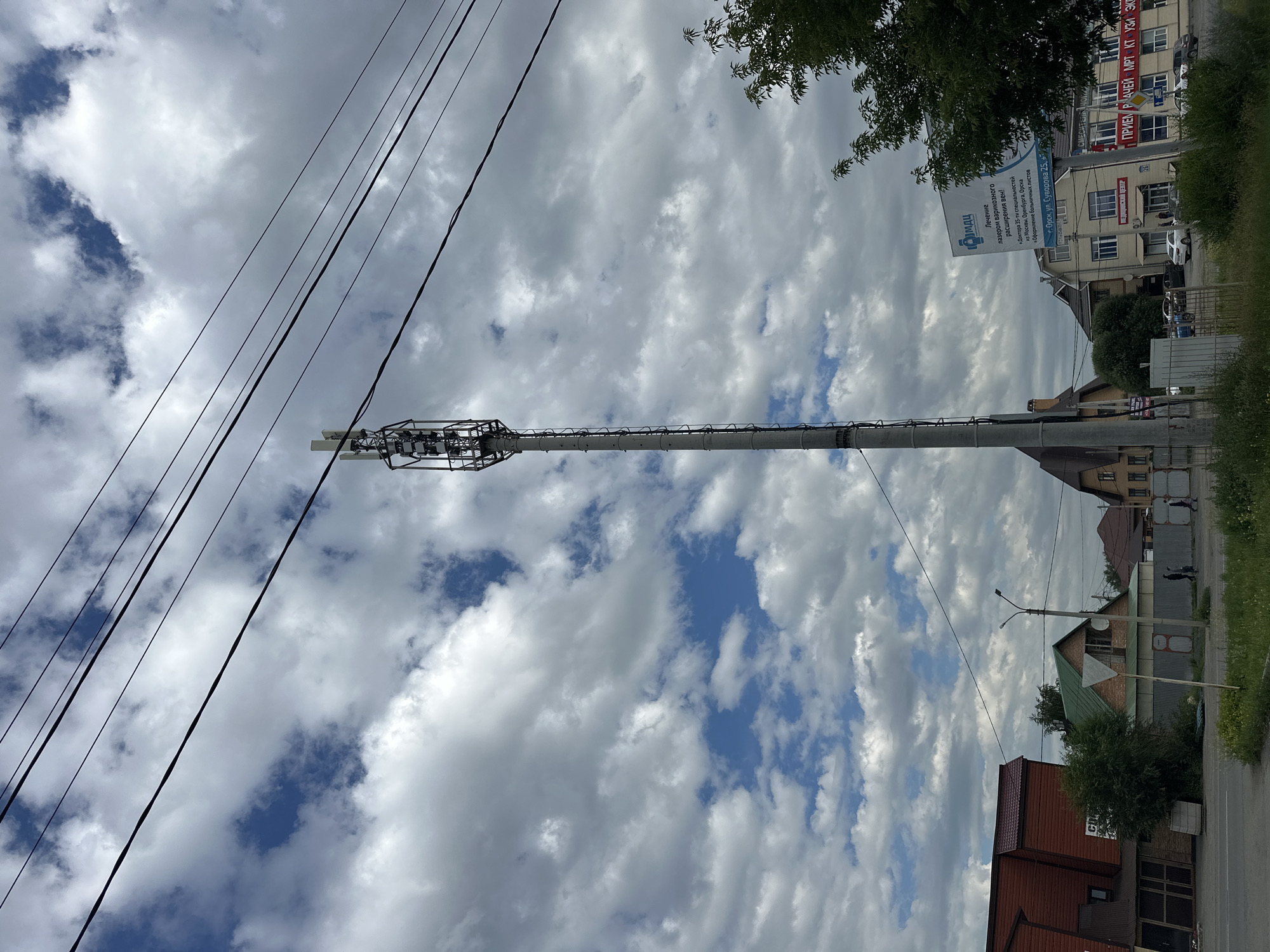 The image shows a view from a street in Orsk, Russia, with a tall cell tower dominating the center of the frame. The sky is filled with dramatic, cloudy formations interspersed with patches of blue. Several power lines stretch horizontally across the upper portion of the image.
To the left, a red building with a dark sloped roof is visible, adjacent to a smaller, darker structure. Further back, a house with a green roof and brickwork stands behind a metal fence. A few trees are scattered throughout the scene, including a dense one in front of the green-roofed house and a larger, leafy tree on the right side of the image.
In the middle ground, a few individuals are walking along a sidewalk. A white billboard with blue text is prominently displayed on the right side of the image, advertising medical services. The billboard's text, partly visible, includes "МЦ", "Орск", and an address. A light pole stands near the billboard.
The foreground consists of a paved road on the left and a grassy area with some weeds and a few yellow wildflowers on the right. The overall impression is a semi-urban environment with a mix of residential and commercial buildings, punctuated by the utilitarian presence of the cell tower and overhead wires.