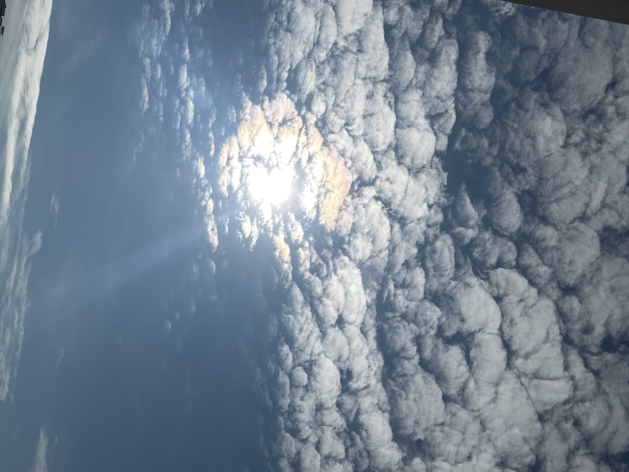 The image shows a sky with patchy, cumulus clouds. The sun is shining brightly, partially obscured by the clouds, creating a radiant effect. Sunlight refracts through the clouds, causing subtle rainbow-like colors to appear in some areas. The sky is a clear blue, indicating fair weather conditions. The bottom portion of the image shows a sliver of a building or structure, suggesting an urban or suburban setting. There are no people or animals visible in the image. The overall scene depicts a bright, partly cloudy day.