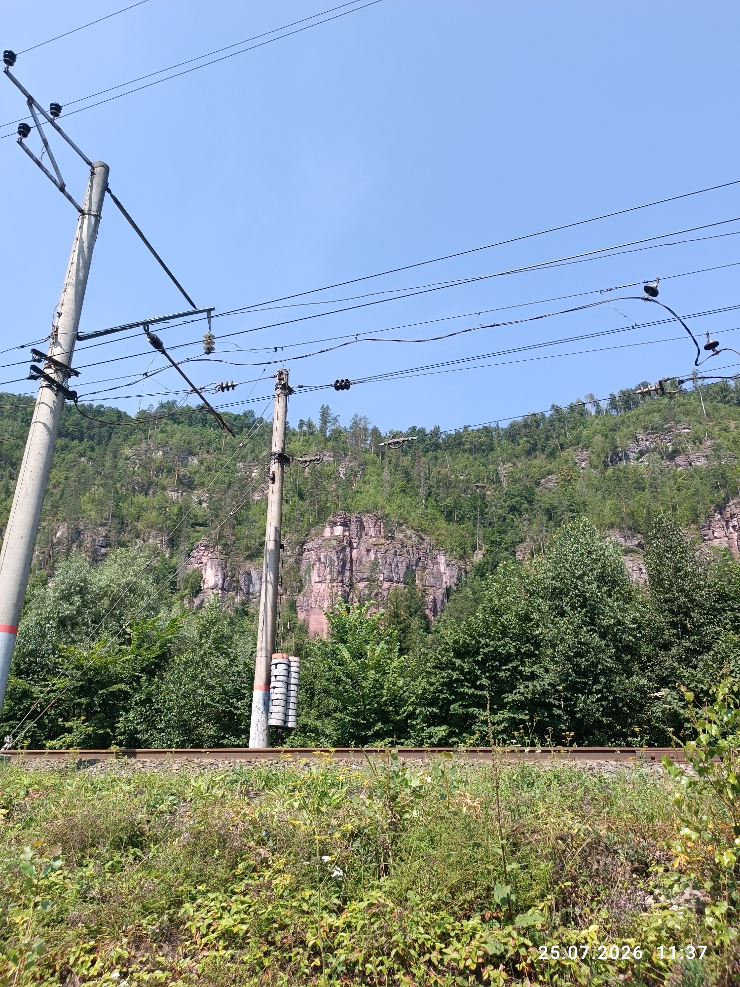 The image shows a clear blue sky over a rocky, tree-covered hillside. In the foreground, there are power lines supported by concrete poles, and a railway track runs horizontally across the frame. Lush green vegetation grows along the sides of the track and the lower slopes of the hill. The date and time stamp "25.07.2026 11:37" is visible in the lower right corner. The scene appears to be a rural or semi-rural area, likely along a railway line, with a natural, hilly landscape.