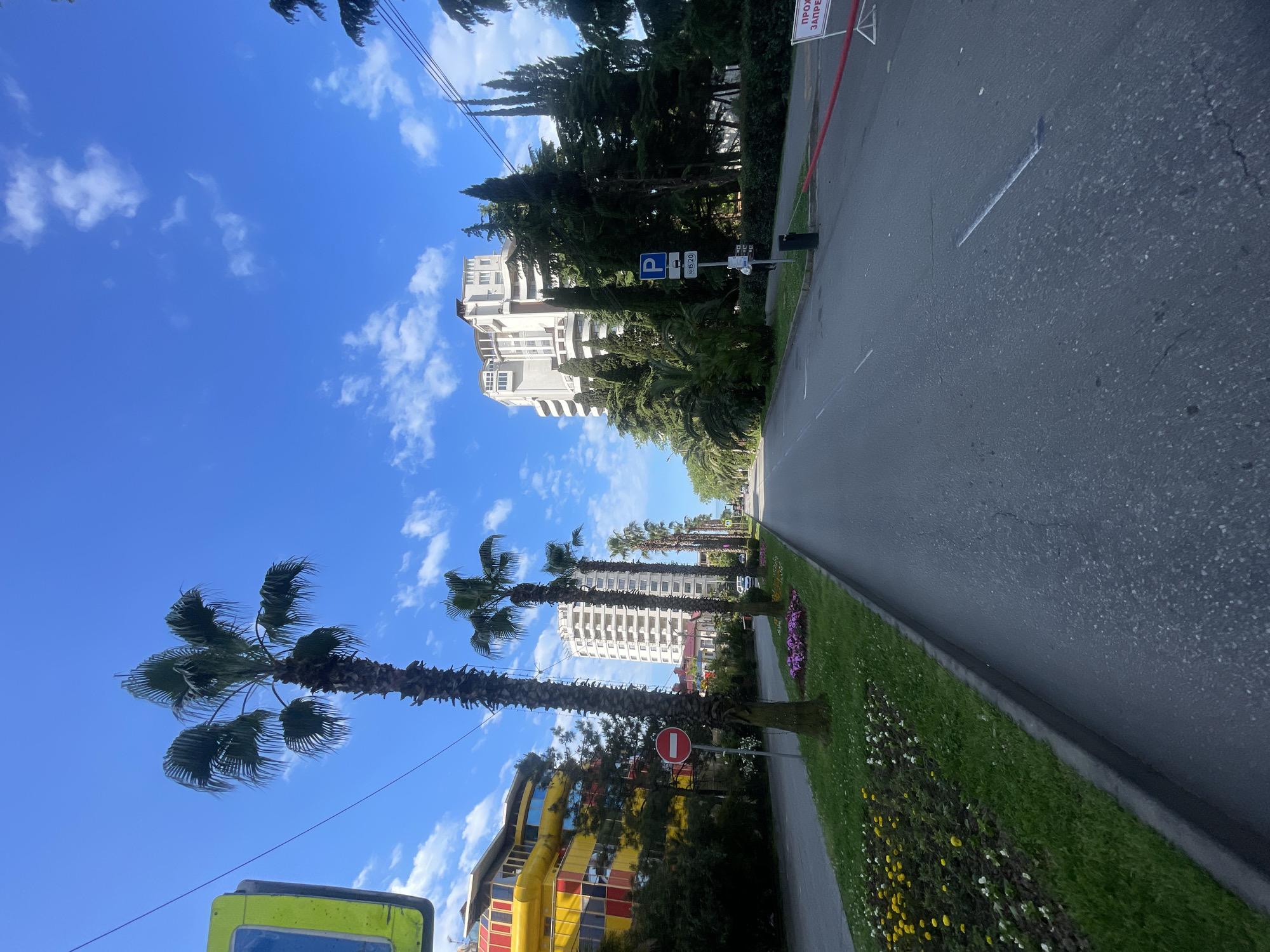 The image shows a street scene in Sochi, Russia. A multi-lane asphalt road runs through the frame, bordered by a green lawn with flowers on the right. On the left are palm trees and buildings, including a tall, white hotel and a brightly colored building with a checkered pattern. A no-entry sign is visible near the checkered building. The sky is blue with scattered clouds. The photograph is taken from a low angle, looking up the street. A parking sign is posted on the right.