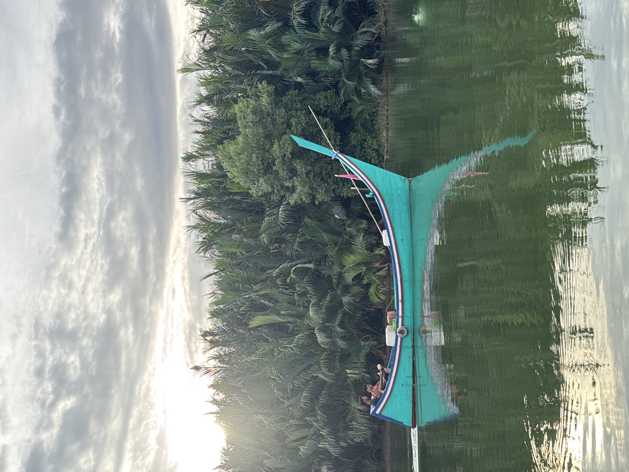 A teal and white boat with two occupants moves through calm green water. The boat has a red and white stripe along its hull. Two young men are seated in the boat, one appearing to be steering. Lush green vegetation, including palm-like trees, lines the shore behind the boat, and the sky is overcast with a hint of sunlight breaking through. The scene reflects on the water's surface. The location is identified as Reudeuep, Indonesia.