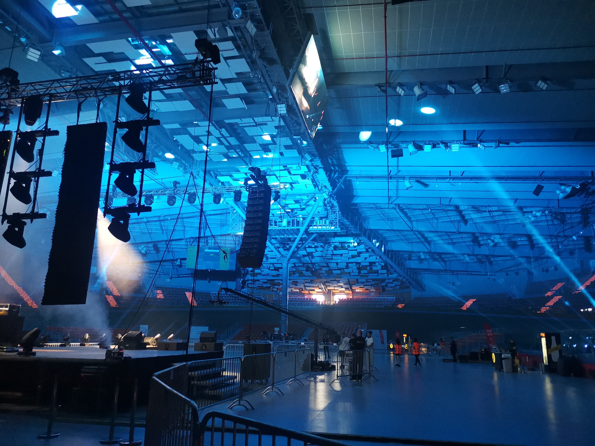 The image depicts the interior of a large, multi-functional indoor arena, identified as Kolodrum Plovdiv, located in Plovdiv, Bulgaria. The scene is dominated by a pervasive blue lighting scheme, emanating from numerous overhead fixtures and stage lights, creating an atmospheric effect.
In the foreground and to the left, a substantial stage setup is visible. This elevated platform is equipped with extensive professional audio-visual equipment, including multiple large, dark-colored line array speaker stacks suspended from overhead trusses. Numerous spotlights and stage lights are mounted on these trusses and positioned on the stage floor, some emitting visible beams through atmospheric haze or smoke. A large, brightly lit rectangular display screen is suspended from the ceiling towards the center-left.
The arena's high ceiling features a grid pattern of structural elements and integrated lighting fixtures. Beyond the stage, tiered seating stands, mostly unoccupied, curve around the periphery of the arena floor. Sections of these stands in the background are illuminated with orange light, forming horizontal stripes.
On the polished floor of the arena, several metal crowd barriers are arranged, delineating areas. Multiple individuals, appearing to be event staff or crew, are present on the floor, particularly in the mid-ground towards the right. Some individuals are wearing high-visibility orange vests. A camera jib or crane extends from near the stage towards the center of the arena floor. Further to the right, what appear to be temporary stands or concession points with signage are visible. The overall impression is of an event venue during setup or preparation, prior to the arrival of a large audience.
