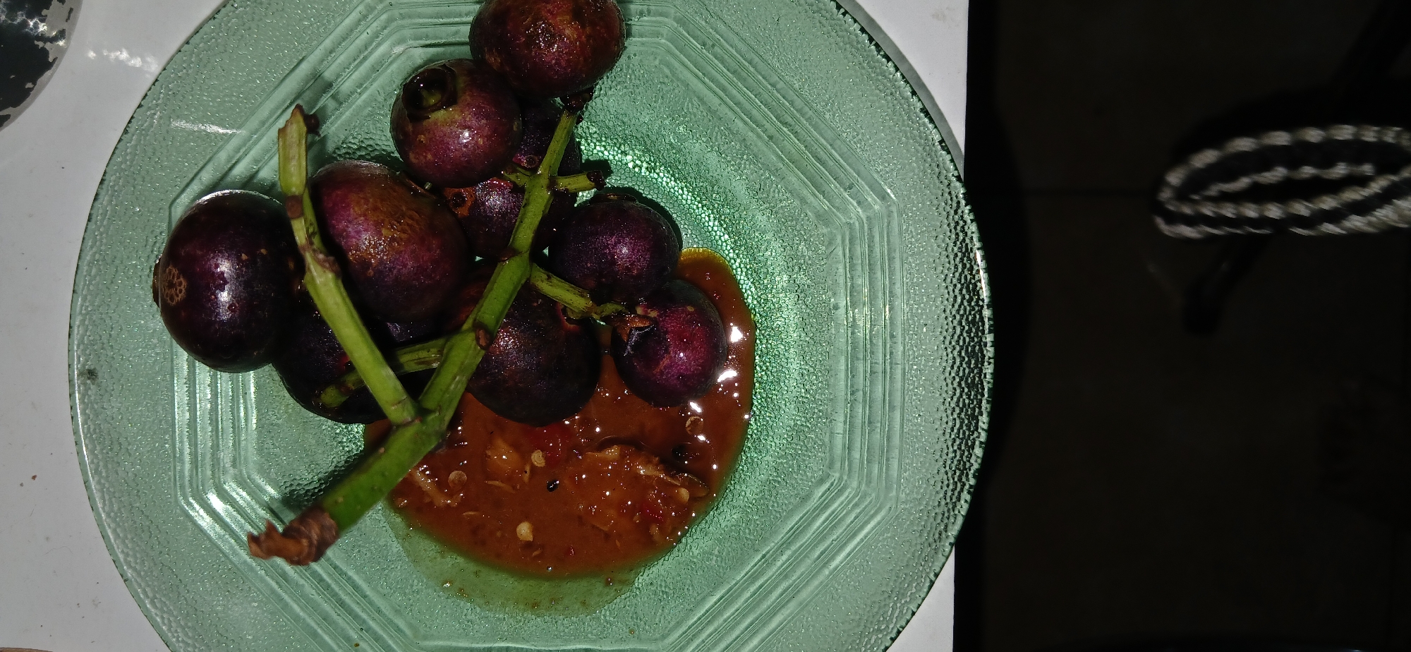 A close-up shot captures a green, octagonal plate on a white surface. The plate contains several dark purple fruits, likely mangosteens, arranged on a cluster of green stems. A small puddle of a reddish-brown sauce, possibly sambal, is to the left of the fruits. The background is out of focus, showing a dark area that may be a table or floor. There are no people visible in the image. The scene appears to be indoors, potentially in a home or restaurant setting in Cigombong, Indonesia.
