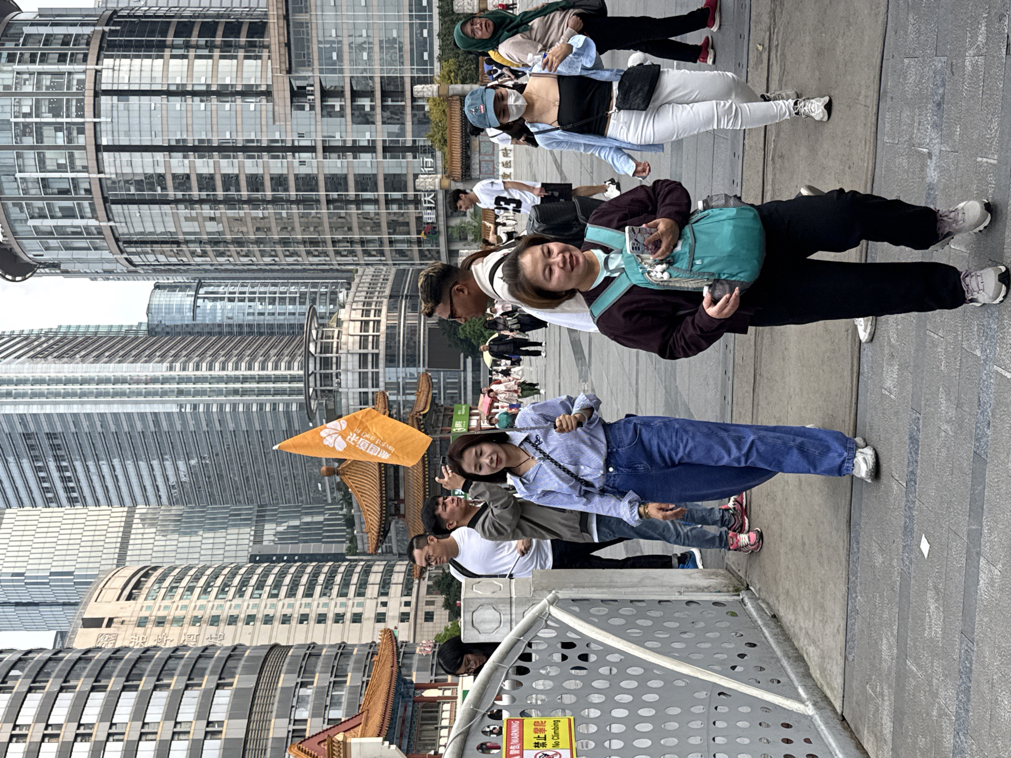 The image captures a bustling street scene in Yuzhong District, China, with a mix of modern skyscrapers and traditional architectural elements. A group of pedestrians are walking, with several people posing for photos. In the foreground, a woman with a turquoise backpack and a dark jacket is smiling at the camera. Behind her is a crowd of people walking. To her left is a group of people, including a woman in a striped shirt and jeans, and another man carrying an orange flag with Chinese characters. The street is paved with gray stone. In the background, a mix of modern buildings and traditional Chinese architecture can be seen.