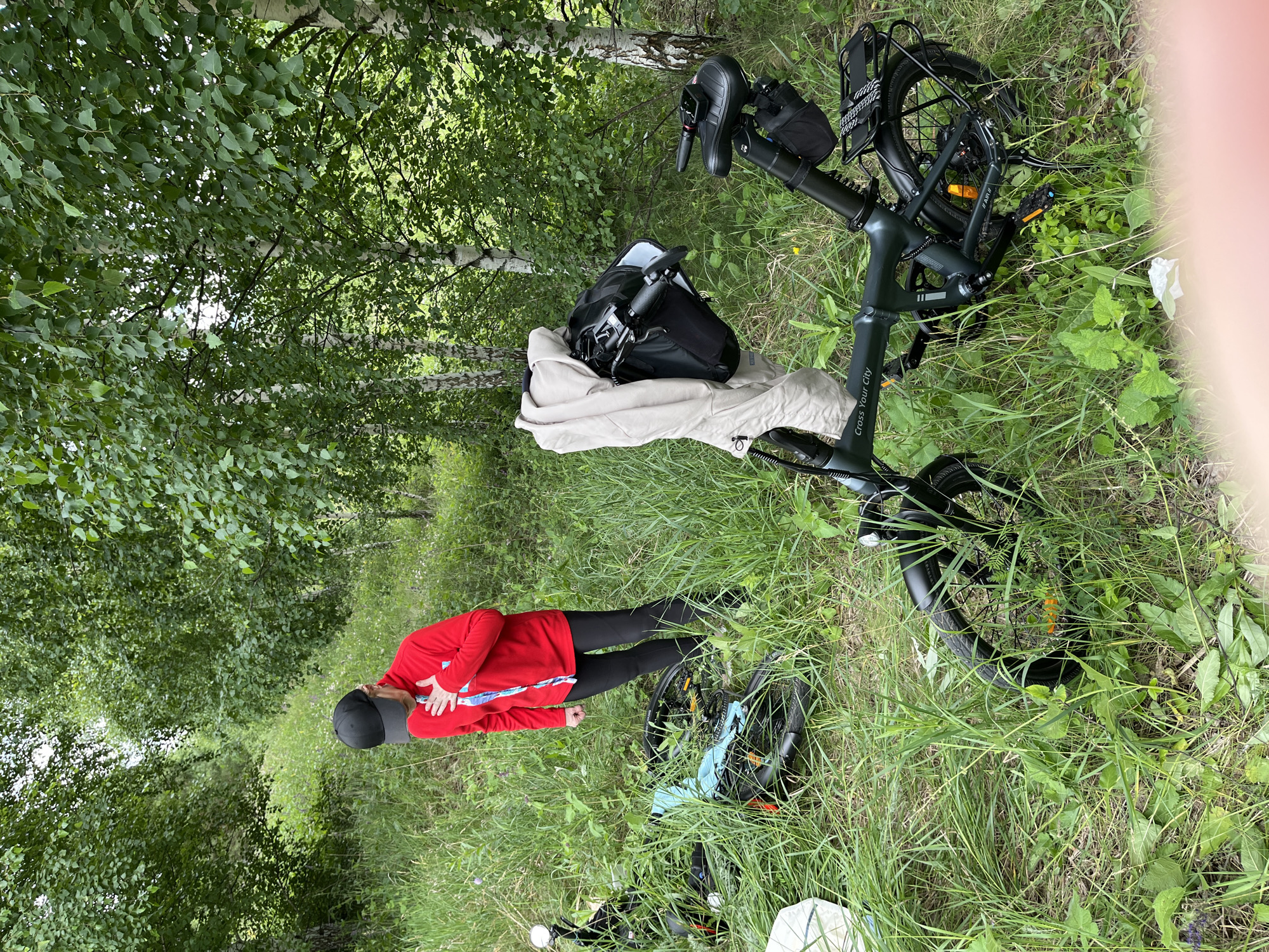 A person in a red long-sleeved shirt and black pants stands on a grassy path next to a folded black electric bicycle. The person is wearing a black baseball cap and is looking down at the bicycle. The bicycle has a black backpack attached to the handlebars and a light-colored blanket draped over the seat. The scene is set in a wooded area with tall green trees and lush grass. The lighting suggests it is daytime, and the weather appears to be overcast or slightly cloudy. Visible text on the bicycle reads "Cross Your City". The location is identified as Новое Надырово, Russia.