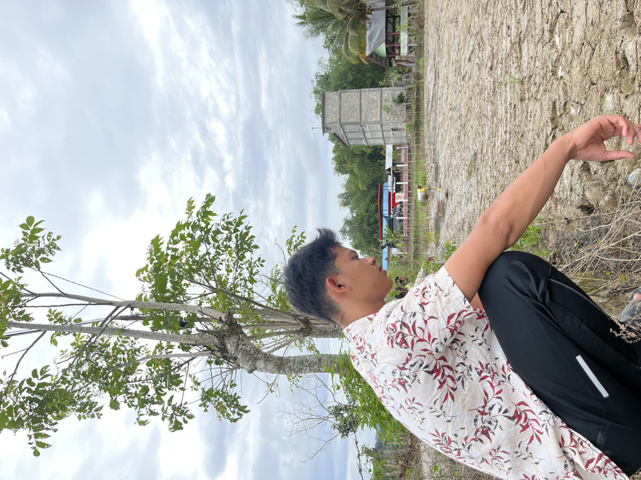 A young man sits outdoors, clad in a floral white shirt and black pants. He is positioned on the ground near dry grass and a small tree with green leaves. A cracked earth surface stretches out behind him. In the distance, a gray concrete building and a small red-roofed structure are visible amidst green trees under an overcast sky. His arm is extended, fingers gesturing towards the landscape.