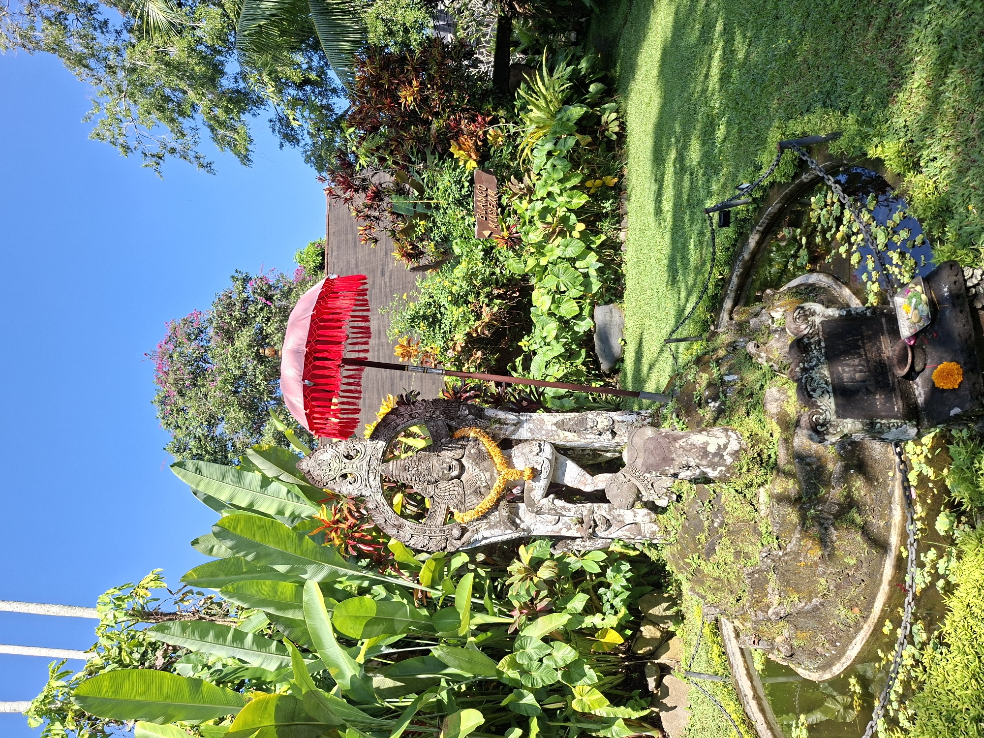 The image shows an outdoor scene in the Ubud District, Indonesia. The primary subject is an ornate stone statue, adorned with a garland of yellow flowers, standing beneath a red umbrella. The statue is surrounded by lush tropical foliage, including banana leaves and various green plants. Behind the statue, there is a garden with a sign indicating "Blanco Renaissance," along with a well-manicured lawn. To the right, a stone water feature, likely a fountain or small pond, is present, partially covered in moss and vegetation. The sky is clear and blue, with trees visible in the background.