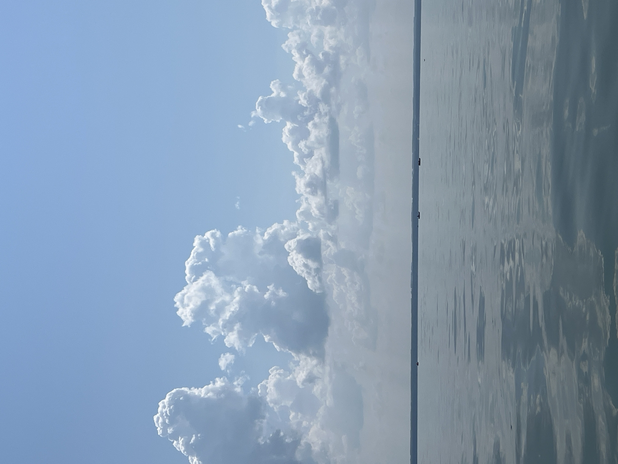The image depicts a serene, wide expanse of water under a bright blue sky dotted with large, fluffy white clouds. The horizon is a faint, thin line, suggesting a vast body of water, possibly a lake or the sea, meeting a distant landmass. The water's surface is calm, reflecting the sky and clouds, with subtle ripples disturbing the mirror-like quality. In the distance, several small boats are visible, scattered across the water. The scene is devoid of any immediate structures or prominent features, creating a sense of openness and tranquility. The description does not contain information about the city of Borovoe, Russia.