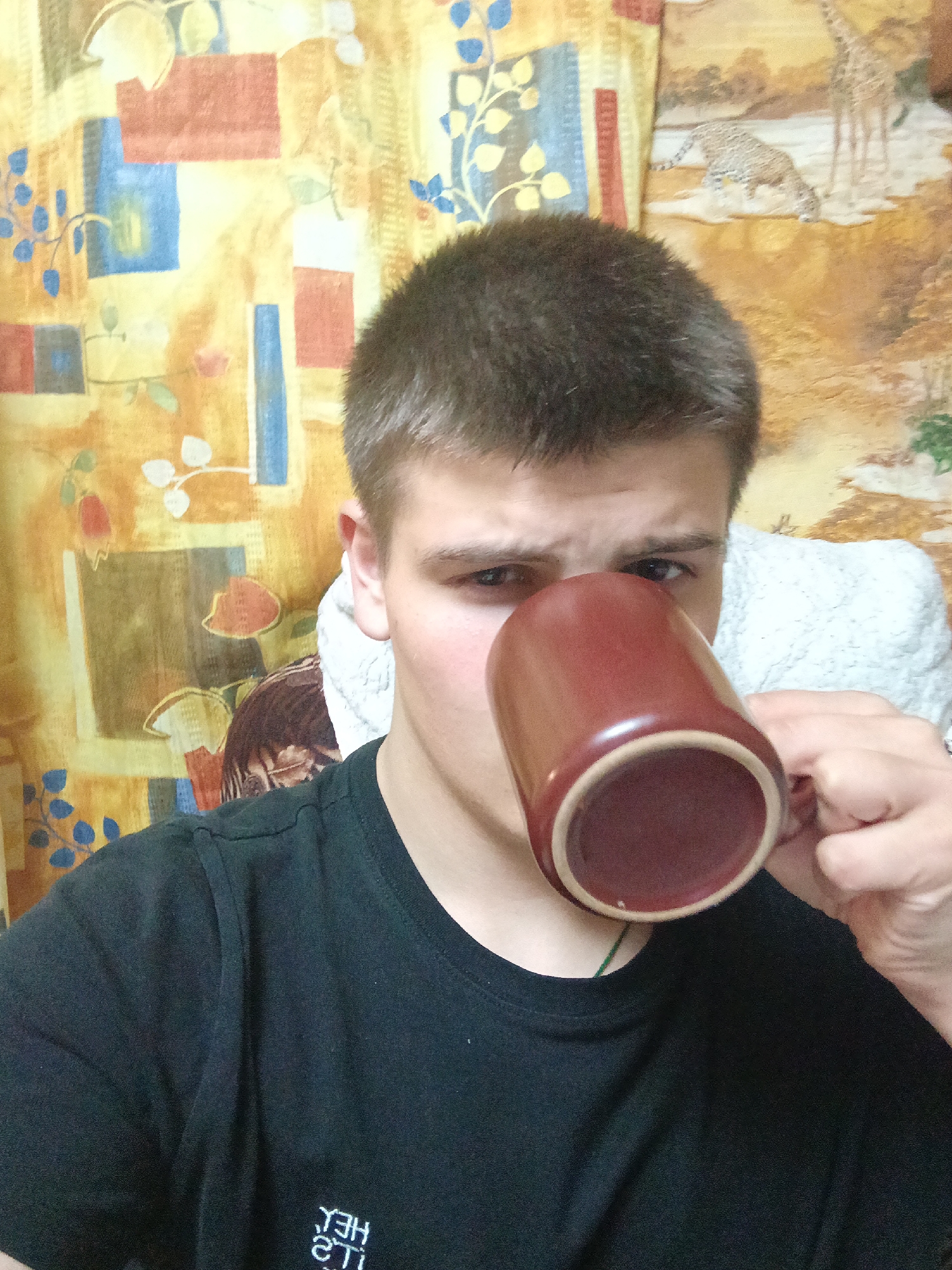 A young man with short brown hair is holding a brown mug with a beige interior up to his mouth. He is wearing a black t-shirt with white lettering at the bottom. Behind him is a patterned tapestry. The background includes what looks like a white cushion or blanket. No details point to Красный Маяк, Russia.