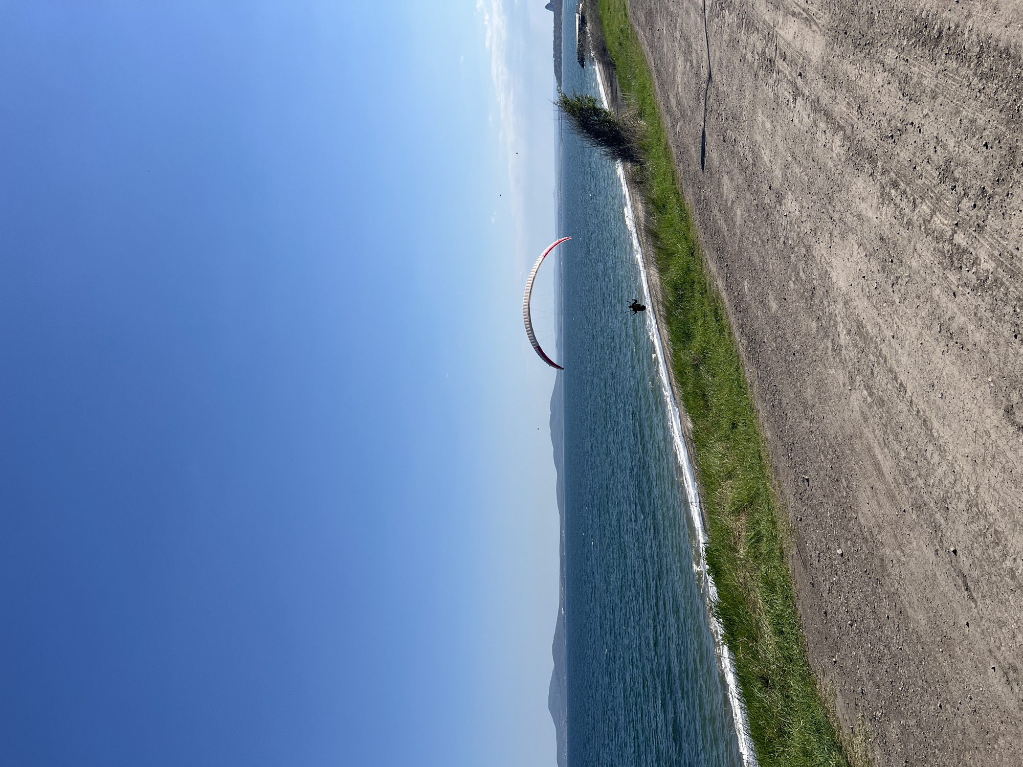 The image shows a paraglider flying over the water near a coastline. The sky is clear blue. The water has small waves and a visible shoreline. The coastline has green grass and a dirt path running alongside the water. The exact location appears to be near Burgas, Bulgaria, based on similar images of paragliding in the area.