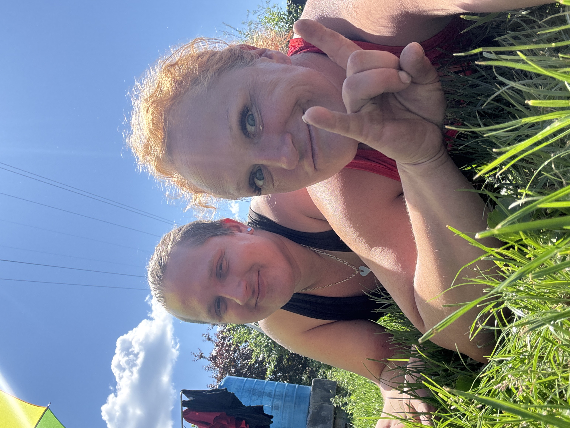 Two individuals are lying in grass outdoors, with a bright blue sky and some clouds above. The person on the left, closer to the foreground, has light brown hair pulled back and is wearing a black tank top with a silver necklace. They are smiling at the camera. The person on the right has bright red hair and is wearing a red tank top. They are looking at the camera with a playful expression and have their right hand raised, making a peace sign. In the background, there's a blue water tank, some laundry hanging on a rack, trees, and power lines stretching across the sky. The overall scene suggests a casual, sunny outdoor setting. The image was taken in the city of Chaikovsky, Russia.