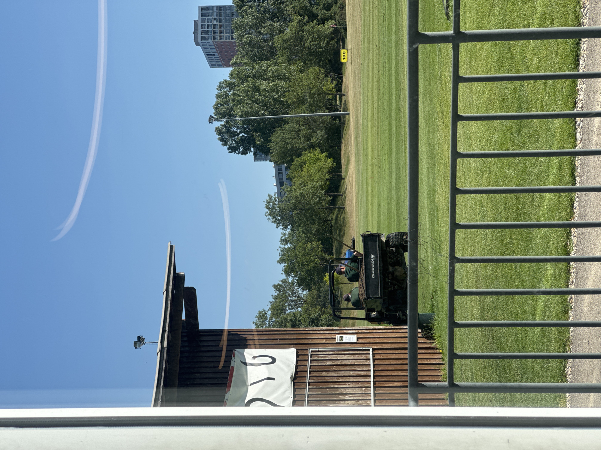 The image depicts a sunny outdoor scene, likely on a golf course or driving range, in Vienna, Austria. In the foreground, a gray metal fence runs horizontally across the bottom of the frame. Behind the fence, a well-maintained green lawn stretches into the distance, with distinct stripes indicating mowing patterns. To the left, a wooden building with vertical planks is visible, featuring a white banner with black markings. Two individuals wearing green shirts and caps are seated in a dark-colored utility vehicle, possibly a Cushman, which is parked on the lawn. The vehicle is filled with dirt. In the middle ground, a tall light pole stands prominently, surrounded by lush green trees. Further in the background, two multi-story buildings, one with a brick facade, rise above the trees. A yellow marker sign with the number "100" is placed on the lawn. The sky is clear blue with faint, wispy white clouds. The scene appears to be a functional area within a park or sports facility, with grounds maintenance activities underway.