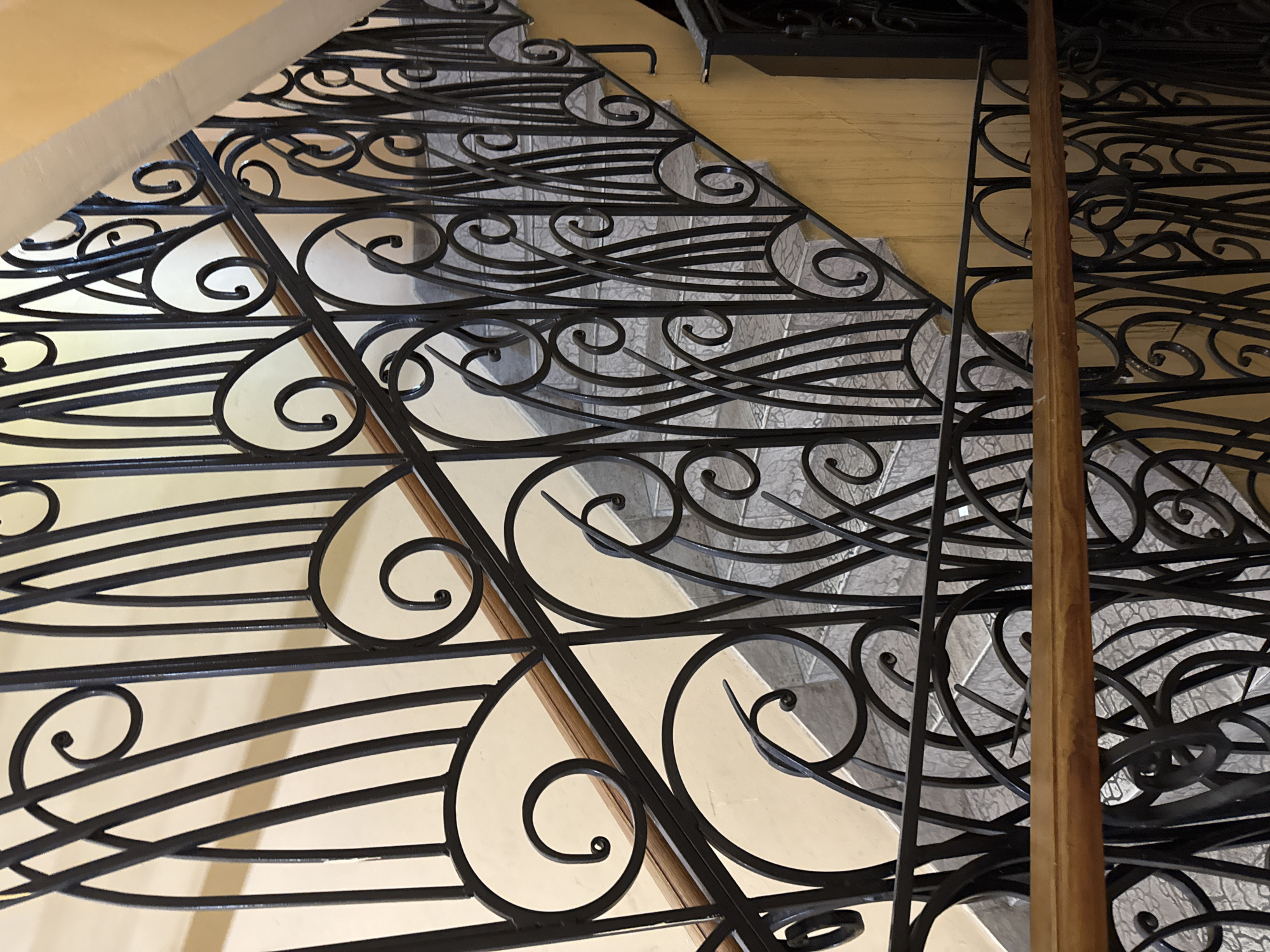 The image shows a close-up view of a black wrought iron railing on a staircase. The railing features intricate scrollwork and curved lines, with a wooden handrail running alongside it. The staircase itself appears to be made of light-colored stone or tile. The setting is indoors, possibly a residential building or a public space. The lighting suggests it is daytime, but there are no external windows or clues to indicate the weather. No people are visible in the image. The focus is entirely on the architectural details of the staircase and railing. The location context is Blagoveshchensk, Russia, but no specific visual cues in the image confirm this. There is no visible text.